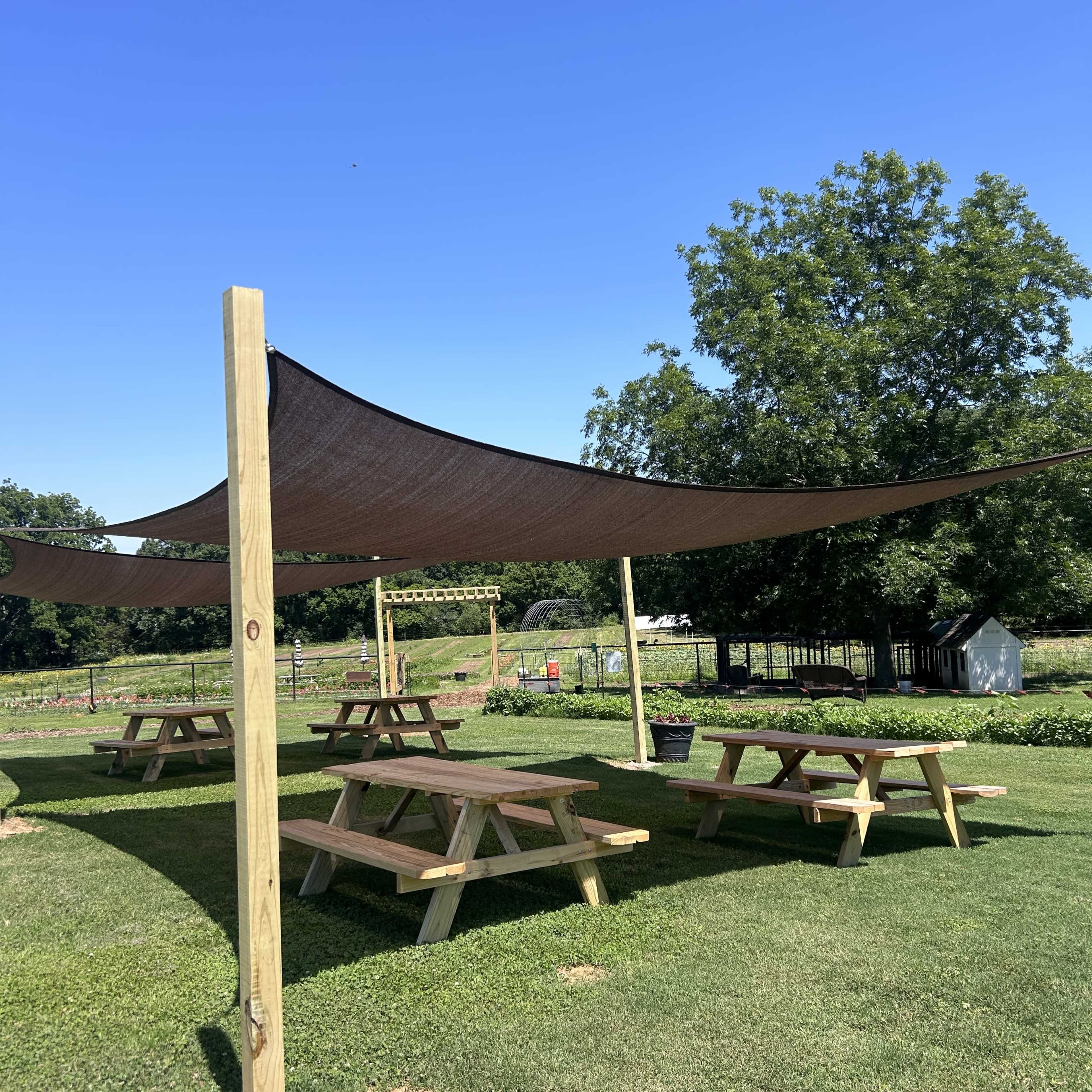 A shaded seating area with picnic tables is set up under a large canopy in a grassy outdoor space.