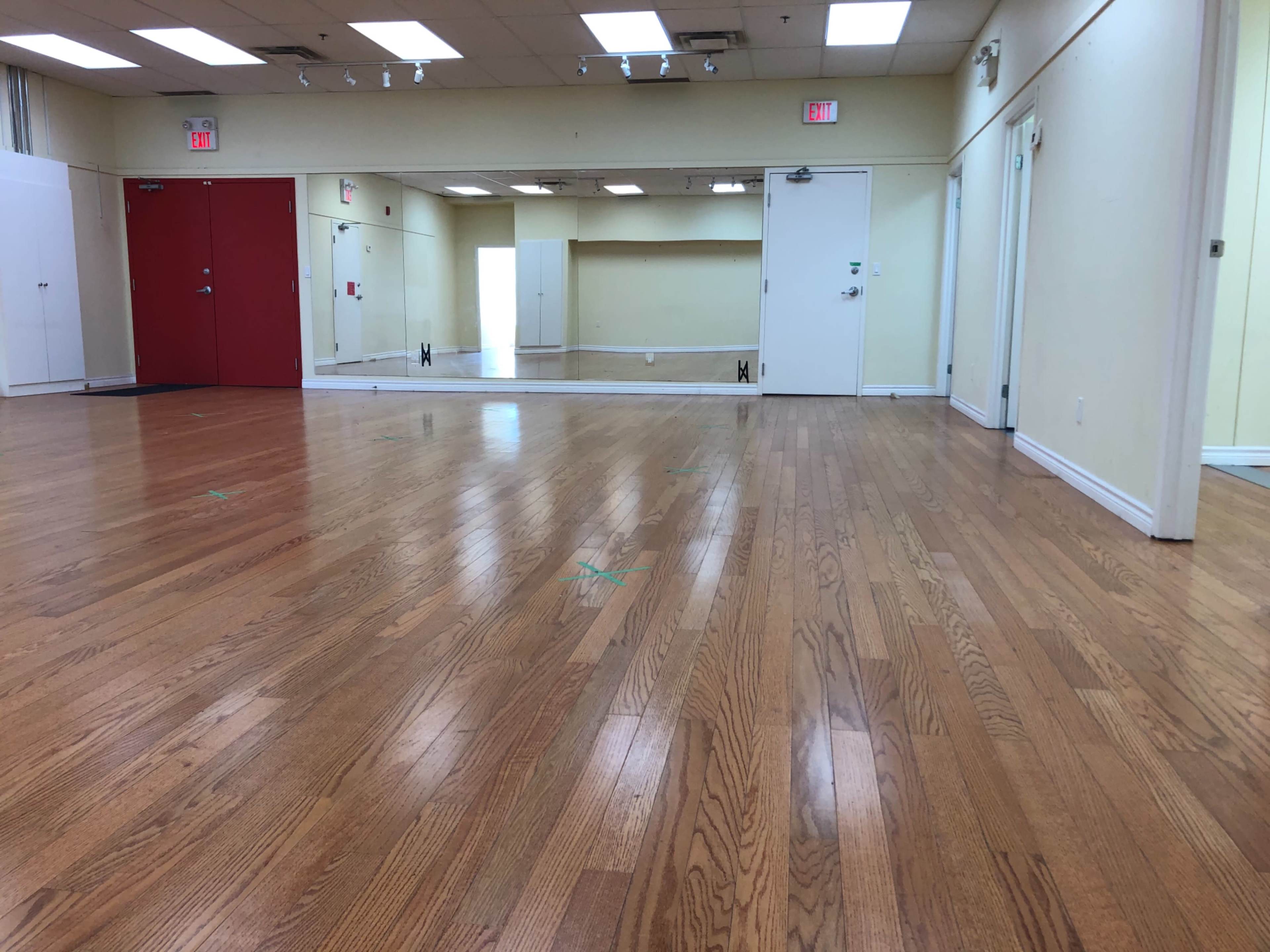 The image shows a brightly lit dance studio with polished wooden floors, two red doors, and large mirrors on one wall.