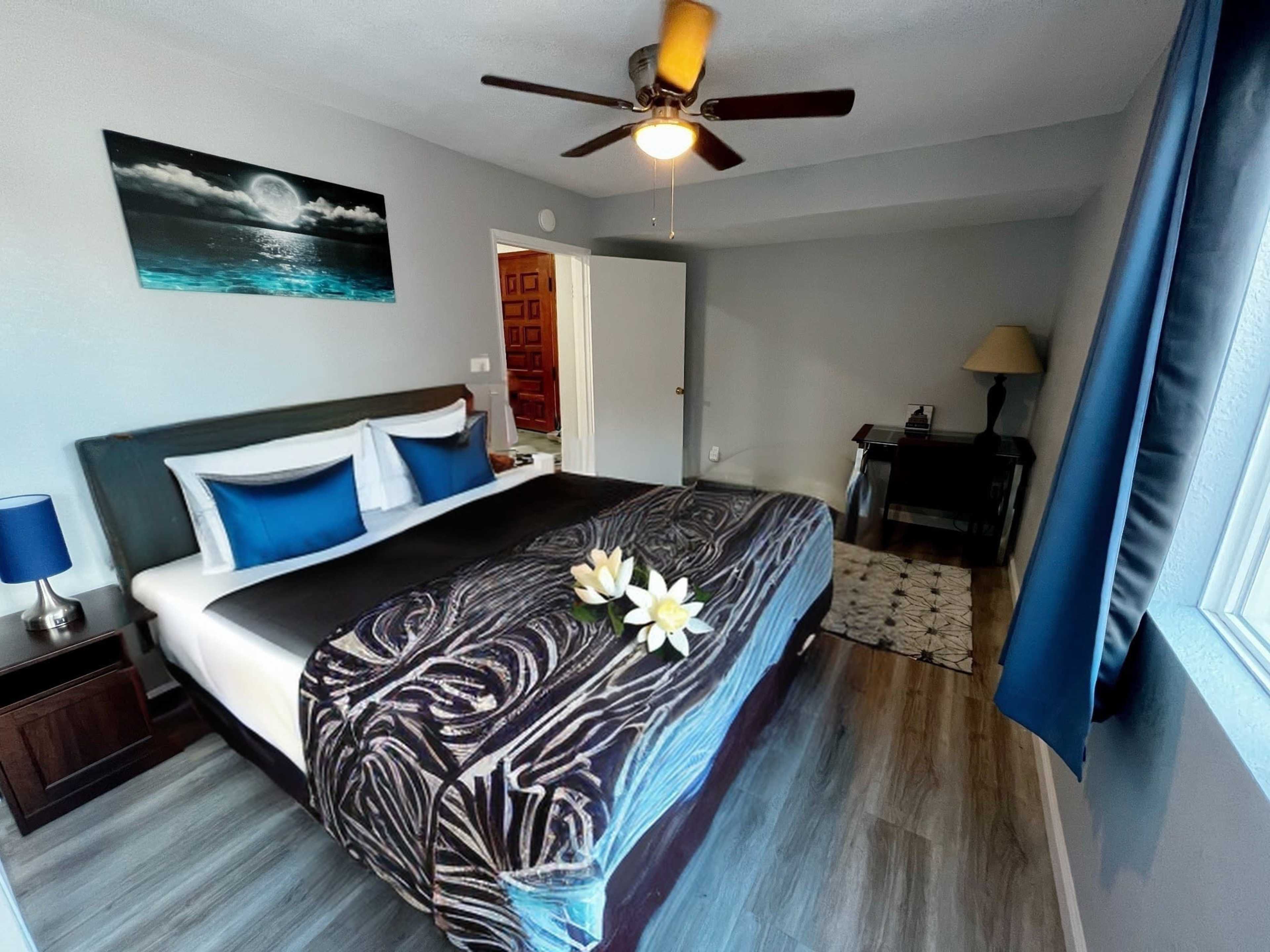 A neatly arranged bedroom features a king-sized bed with blue accent pillows, a ceiling fan, and a small desk with a lamp.
