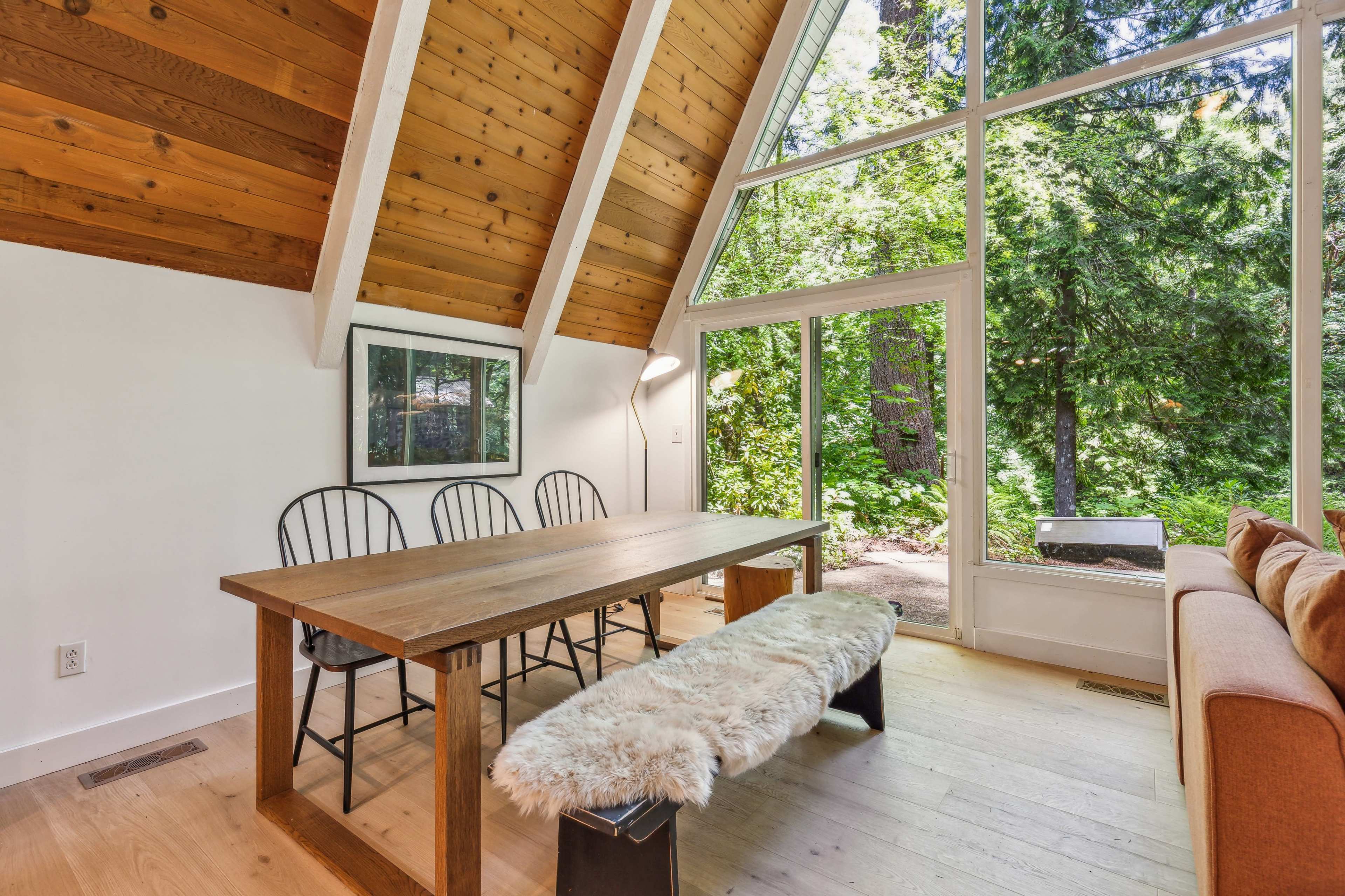 A dining area features a wooden table with three chairs and a bench, set against a backdrop of large windows overlooking a green forest.