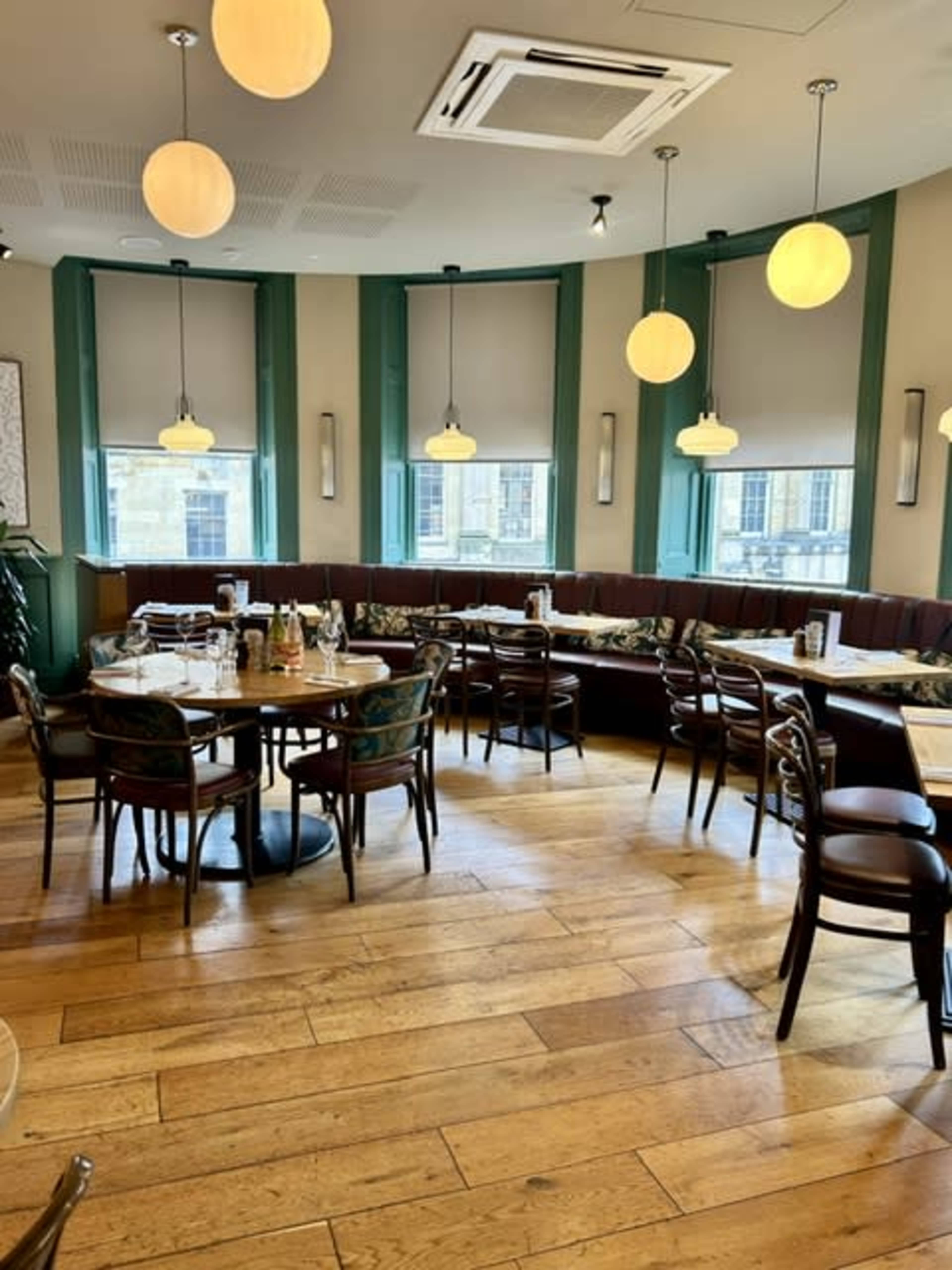 The image shows a restaurant interior featuring wooden flooring, round tables, and a curved booth with green accents and pendant lighting.