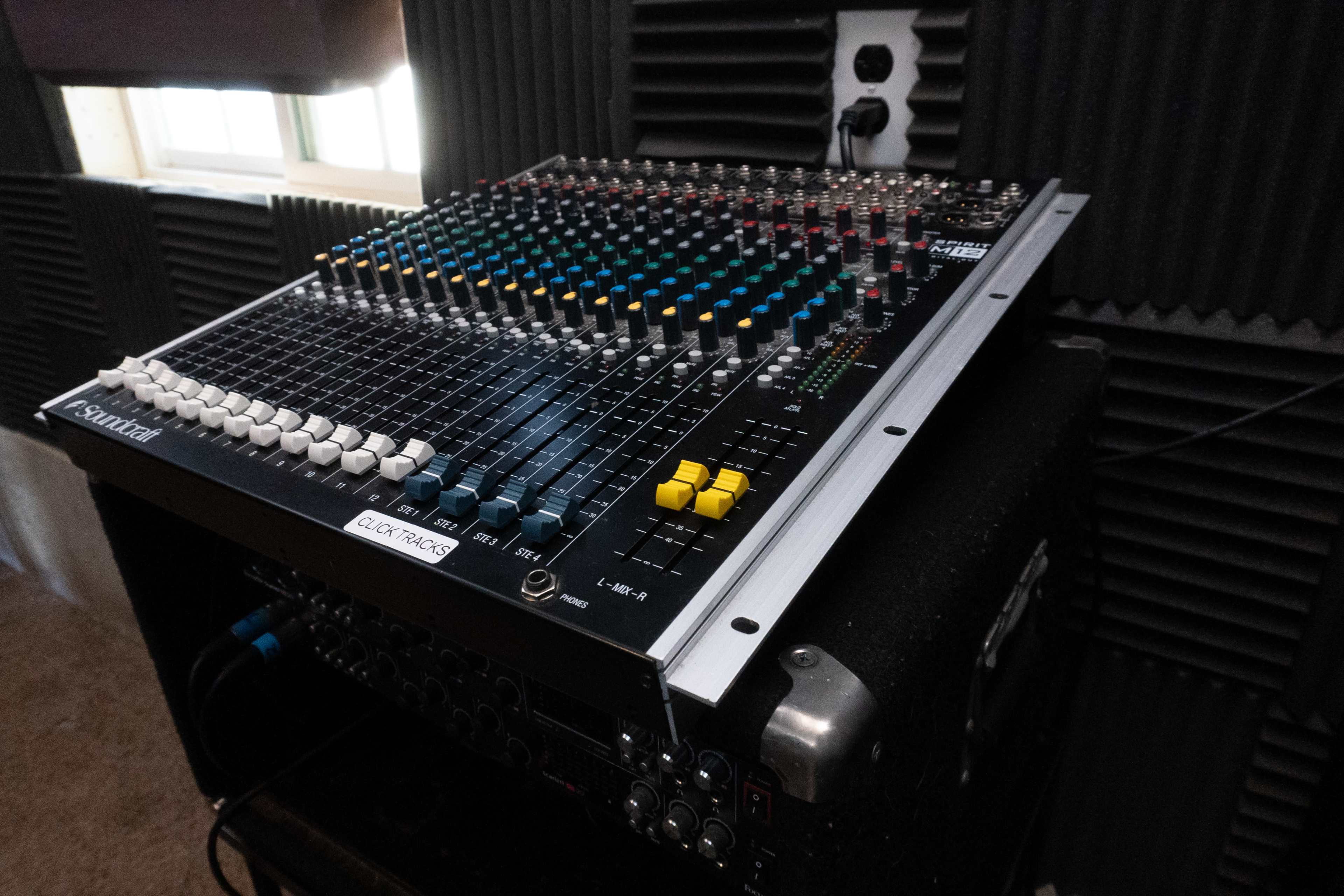 A mixing console with various colored knobs and sliders is positioned on a speaker, surrounded by soundproofing panels.