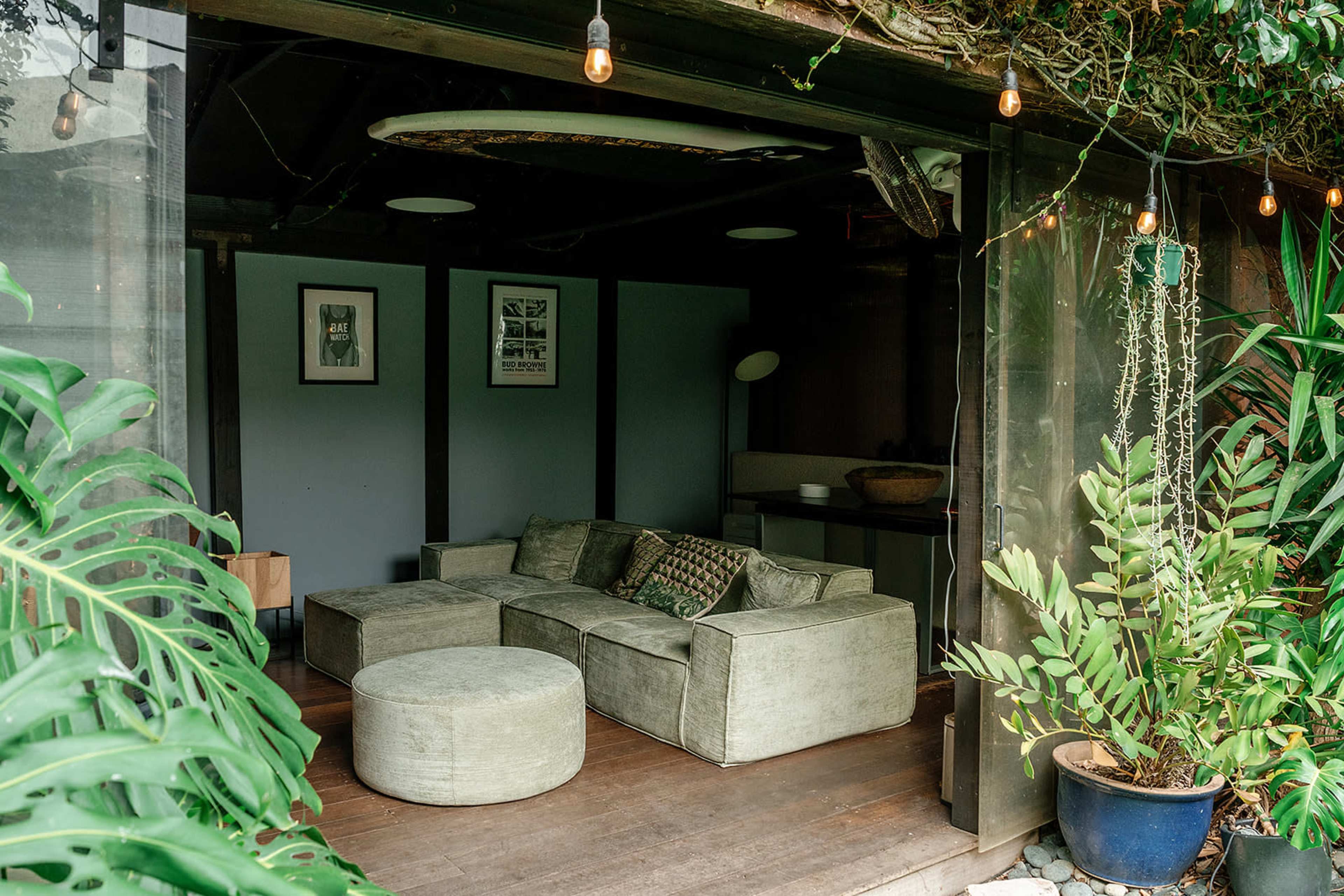 The image shows a cozy living area with a sectional sofa, a round ottoman, and plants surrounding a partially open space.