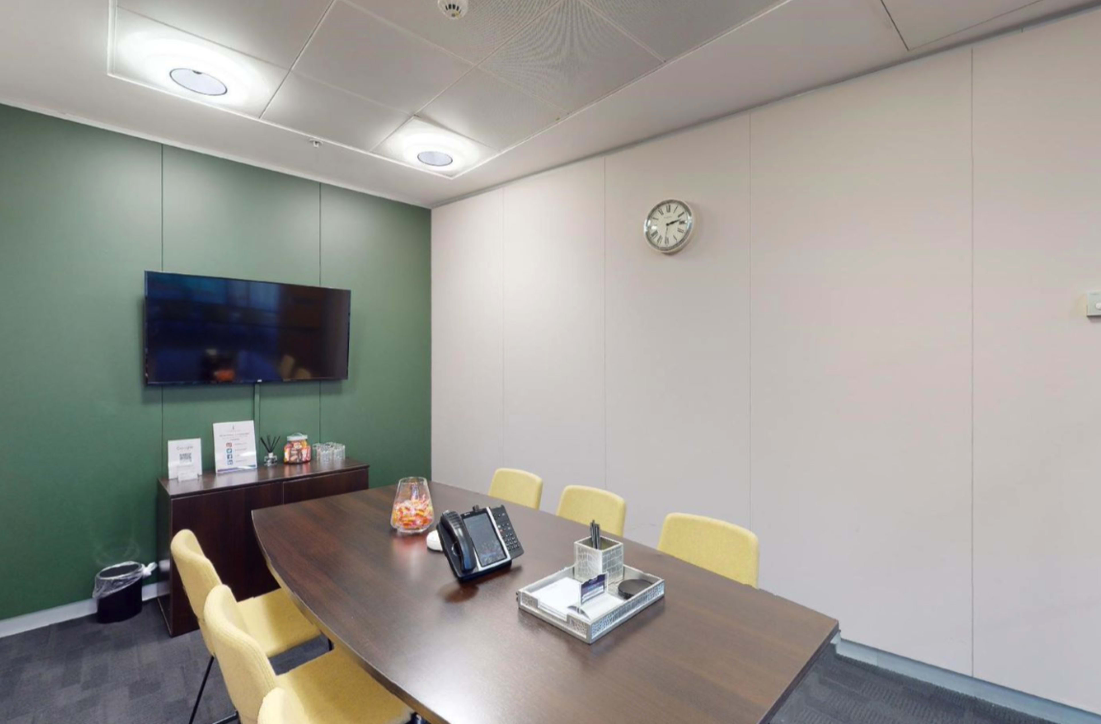 A conference room features a table with yellow chairs, a television mounted on the wall, and a clock above on a plain backdrop.