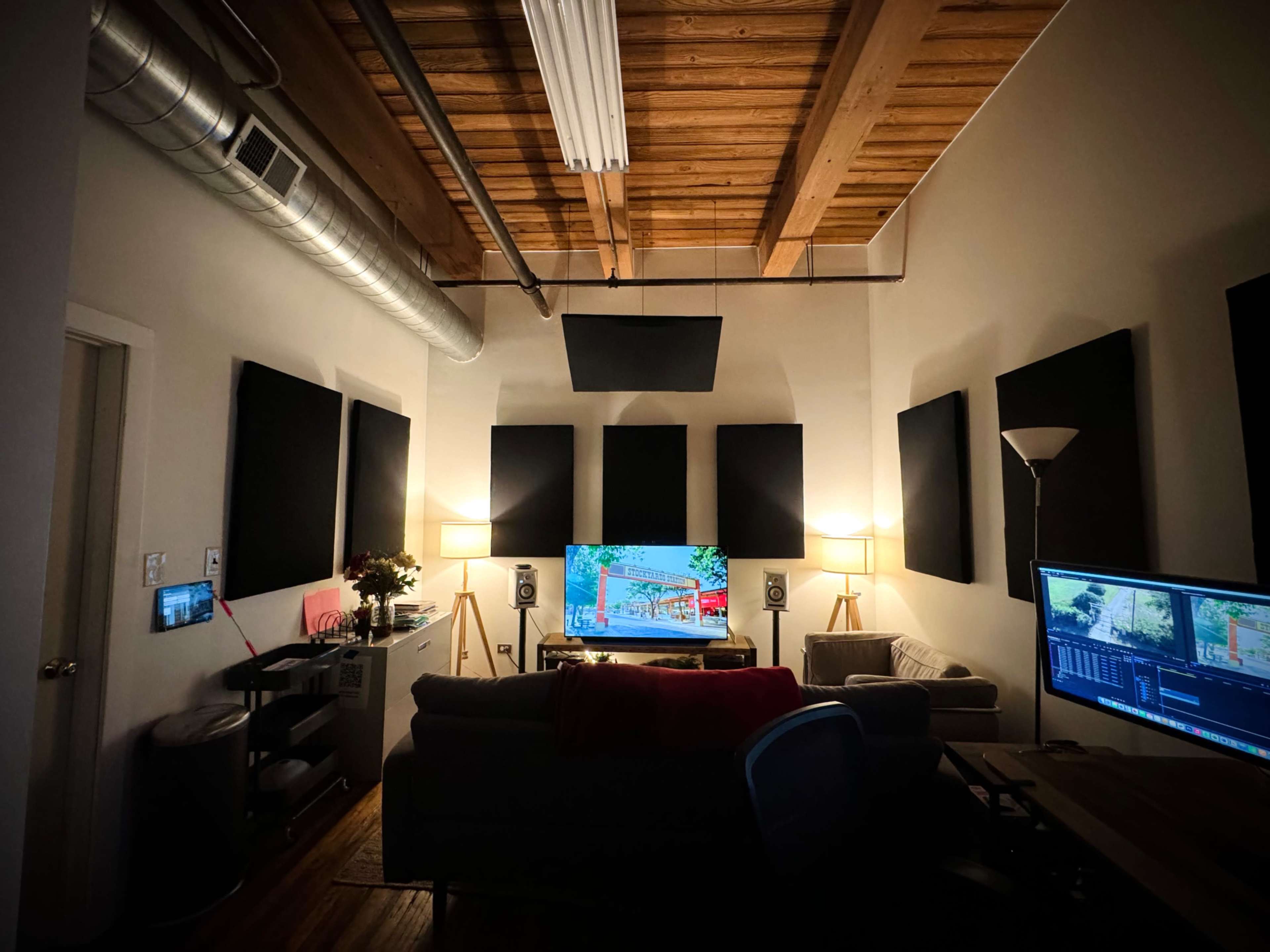 The image shows a dimly lit room with soundproofing panels on the walls, a large screen displaying a colorful image, and two computers on a desk, surrounded by a couch and a floor lamp.