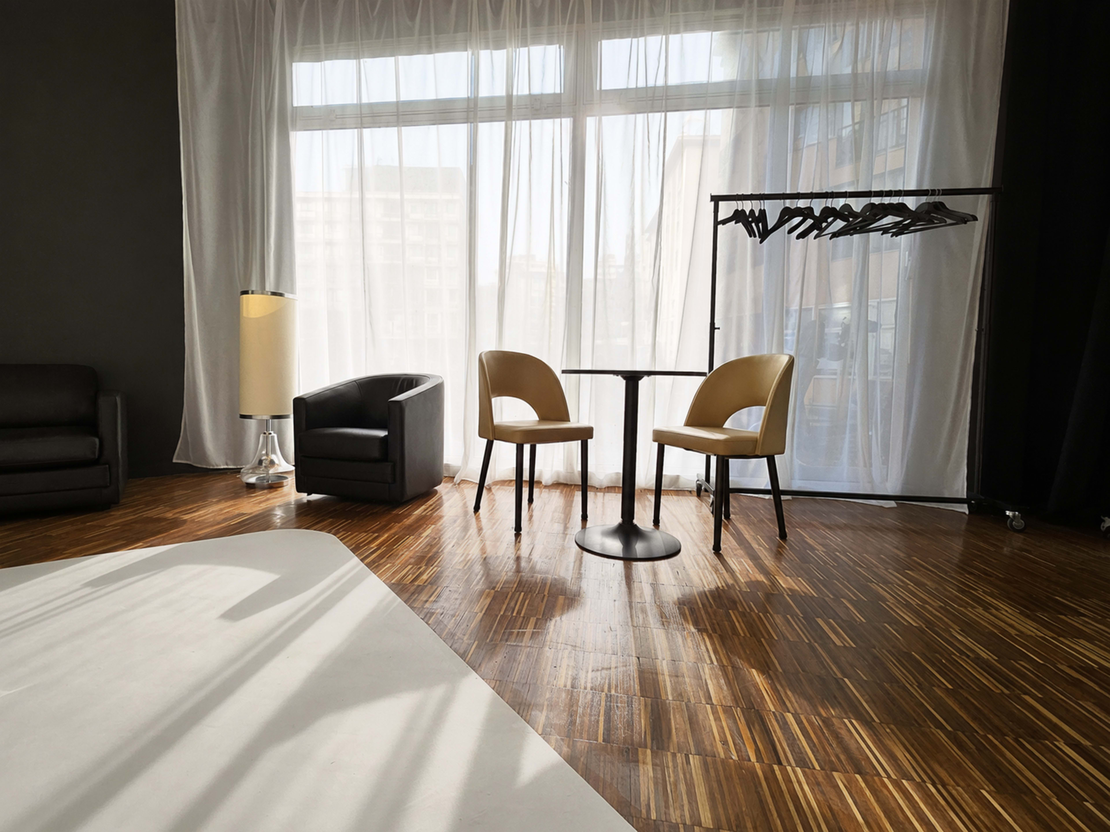 The scene features a well-lit room with two chairs and a small round table, alongside a clothing rack and a lamp, all set against a backdrop of large sheer curtains.