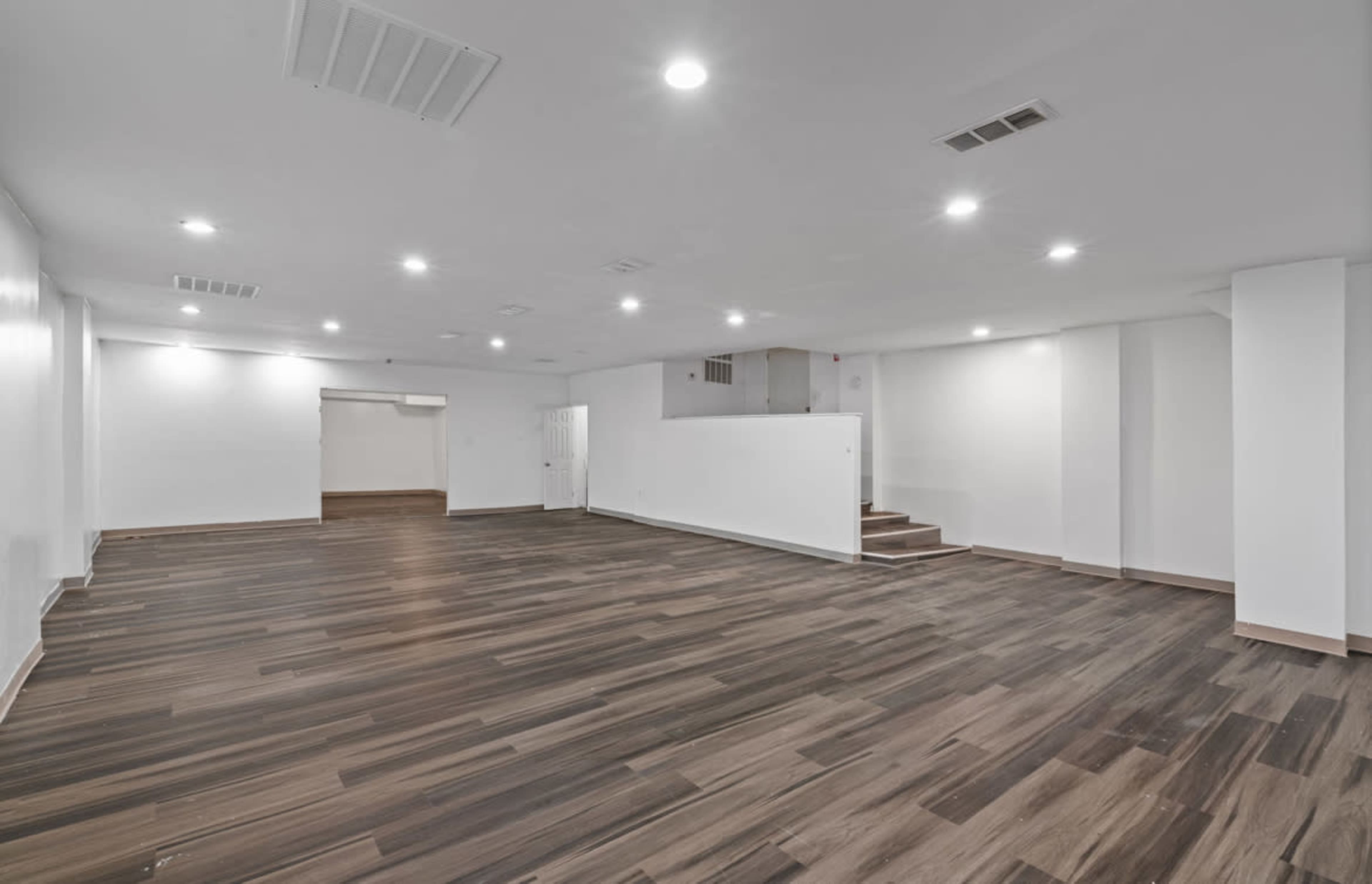 The image shows a spacious, empty room with wooden flooring and bright overhead lighting.