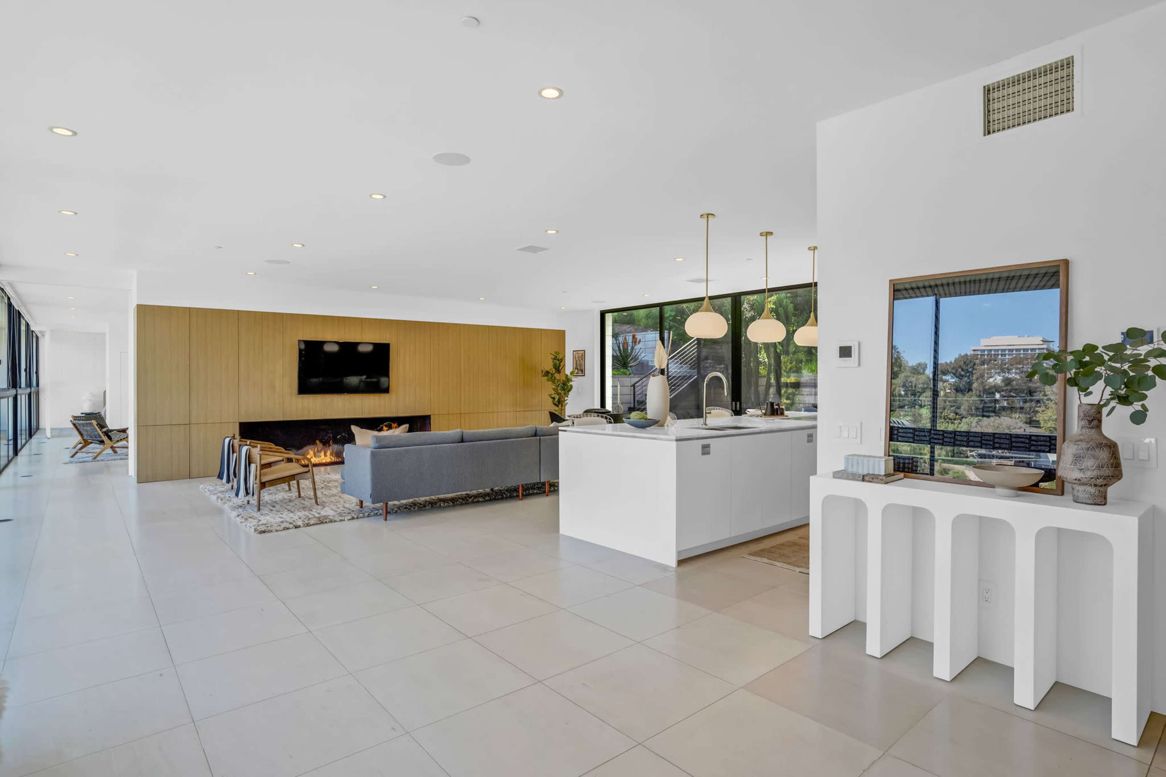 A modern living space features an open layout with a sleek kitchen area, a fireplace, large windows, and minimalist furniture.