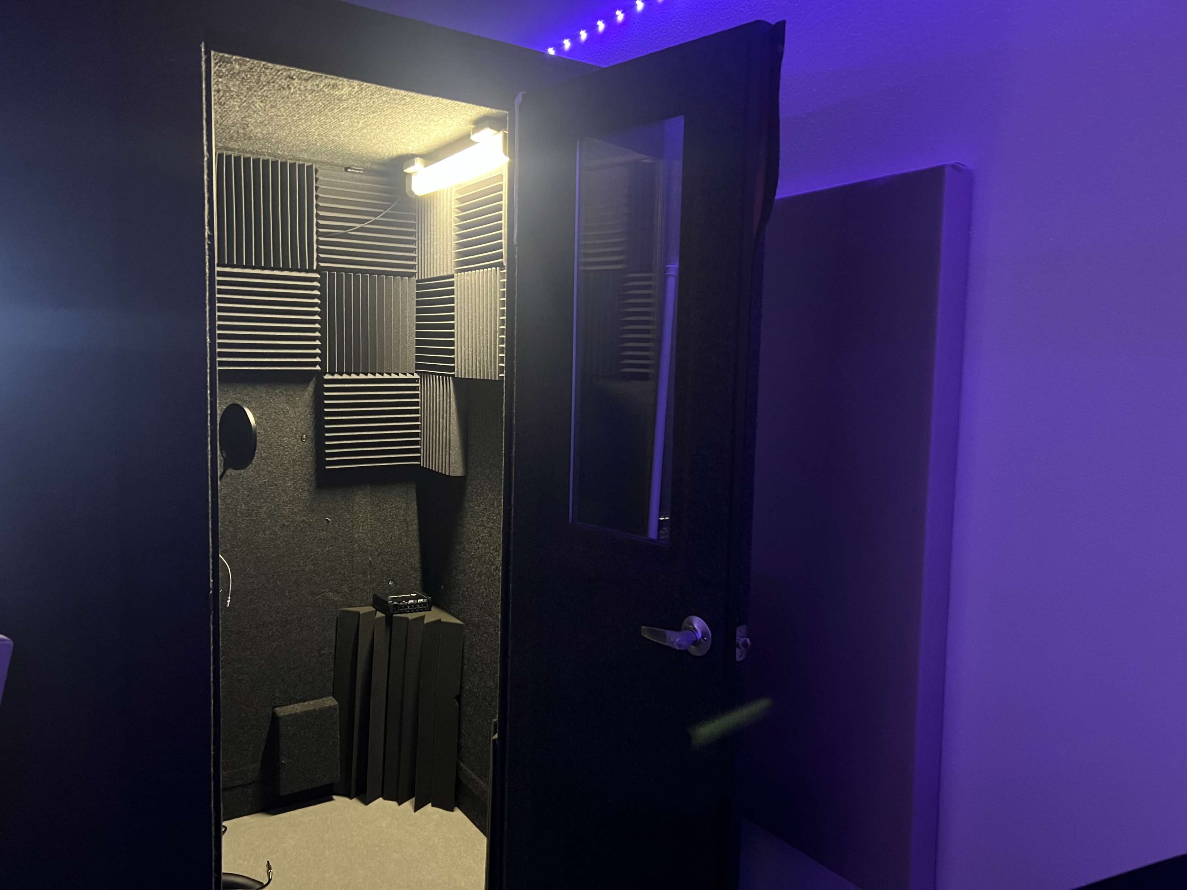 A small soundproof booth with foam panels lining the walls and a microphone stand inside, illuminated by a ceiling light.