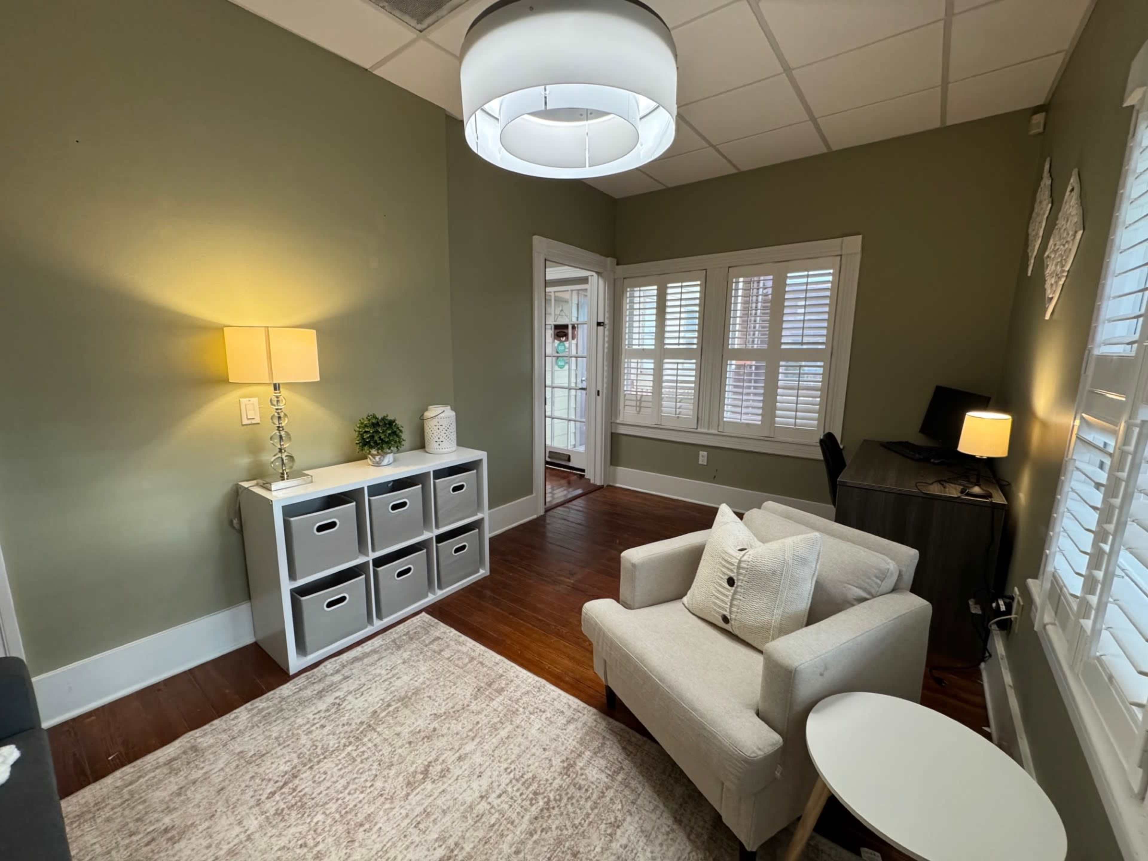 The image shows a small, well-lit room with green walls, a beige armchair, a light fixture, a table, a storage unit with gray bins, and a desk in the corner.