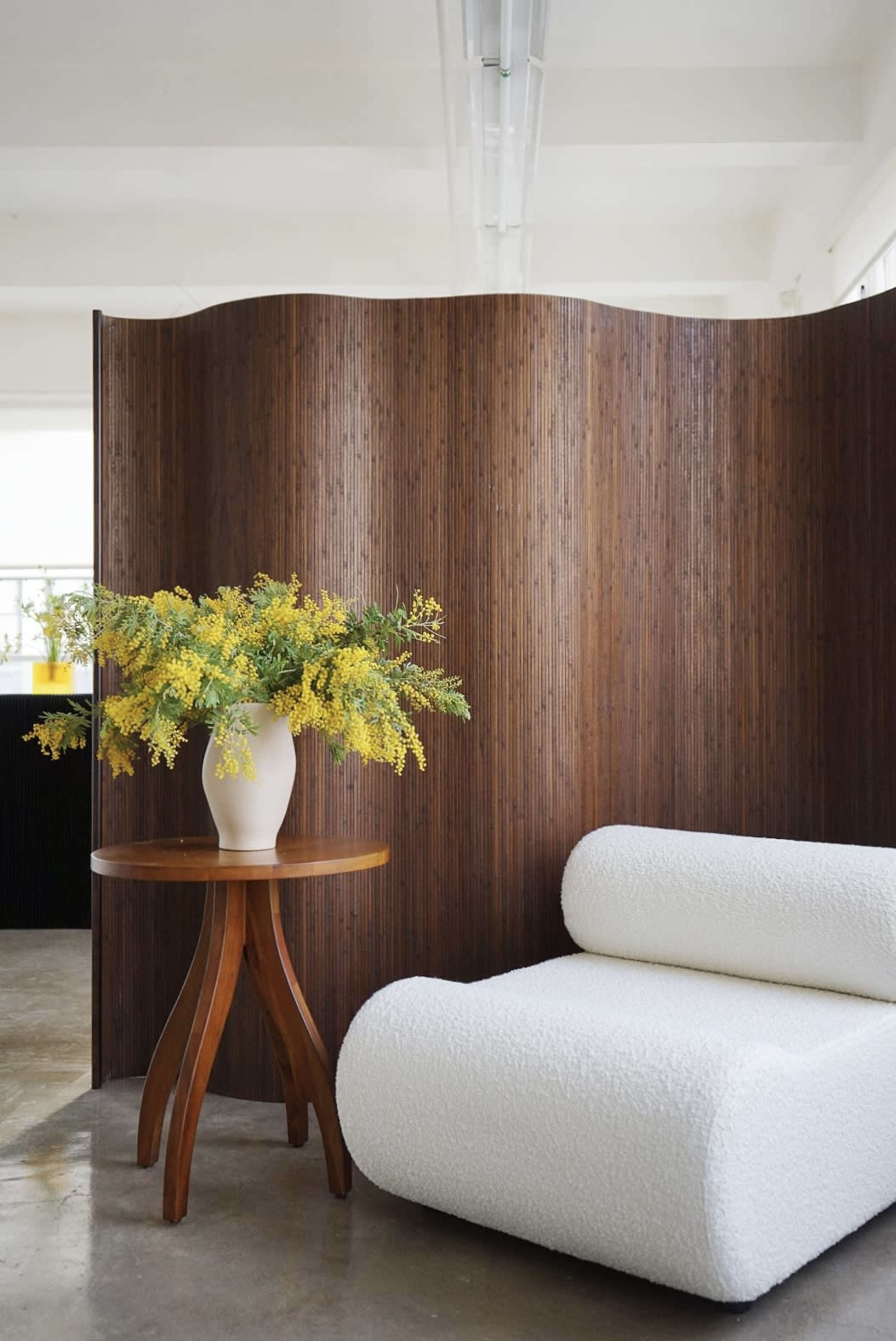 A white, textured lounge chair is positioned beside a wooden table with a vase of yellow flowers, set against a brown wooden screen in a bright room.