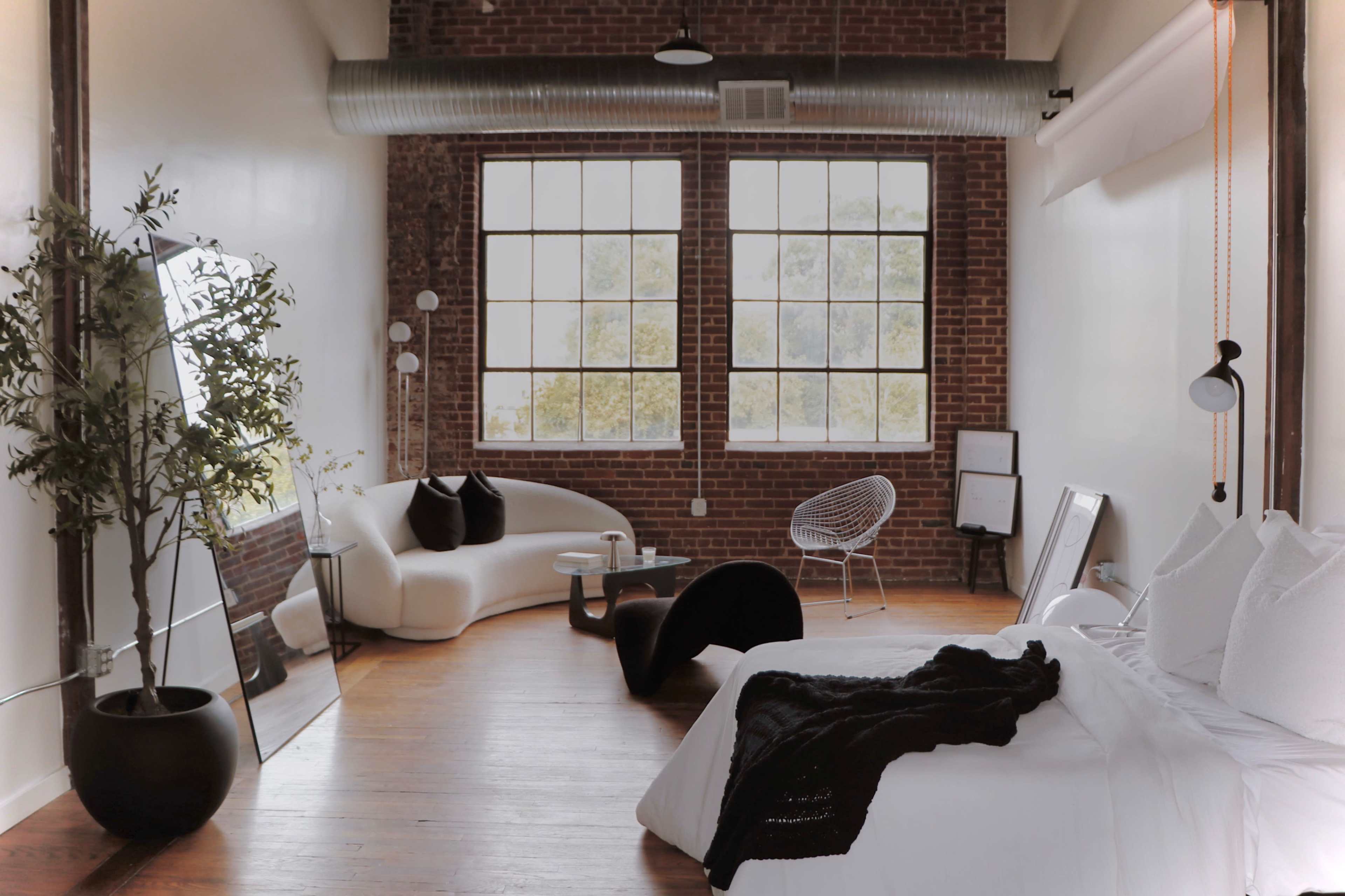 The image shows a modern bedroom with a large bed, a cozy seating area, and exposed brick walls, featuring large windows that let in natural light.