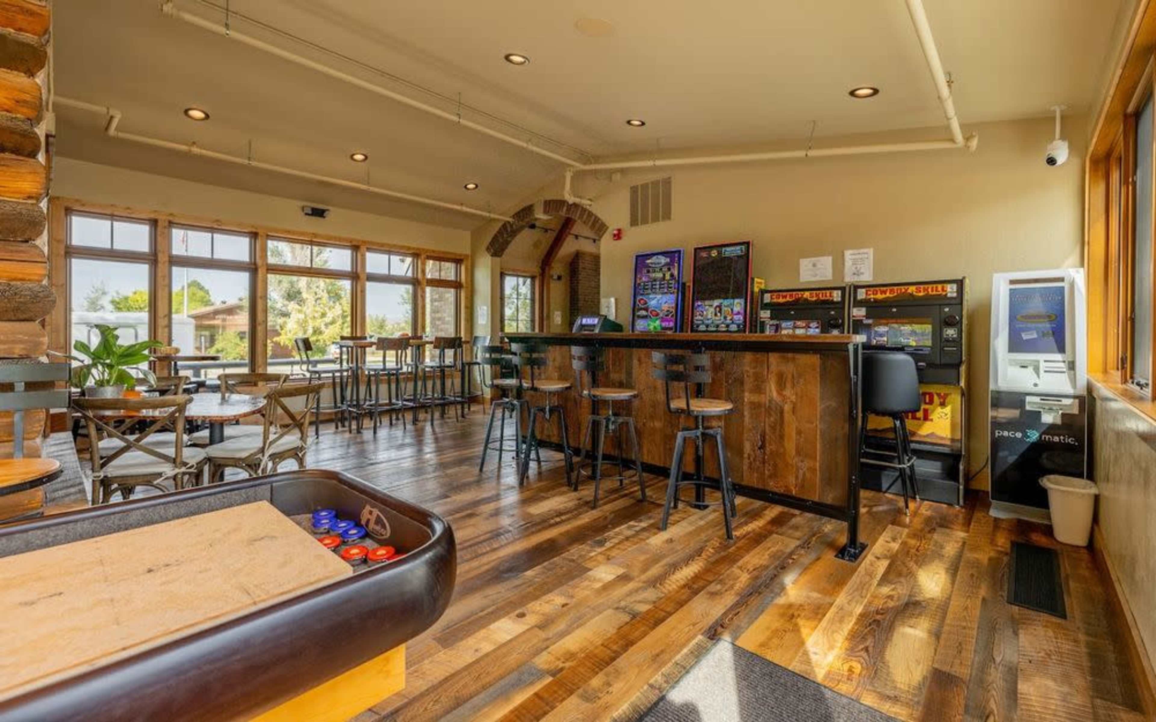 The image shows a spacious, rustic bar area with wooden flooring, a counter with high stools, and multiple slot machines along the walls.