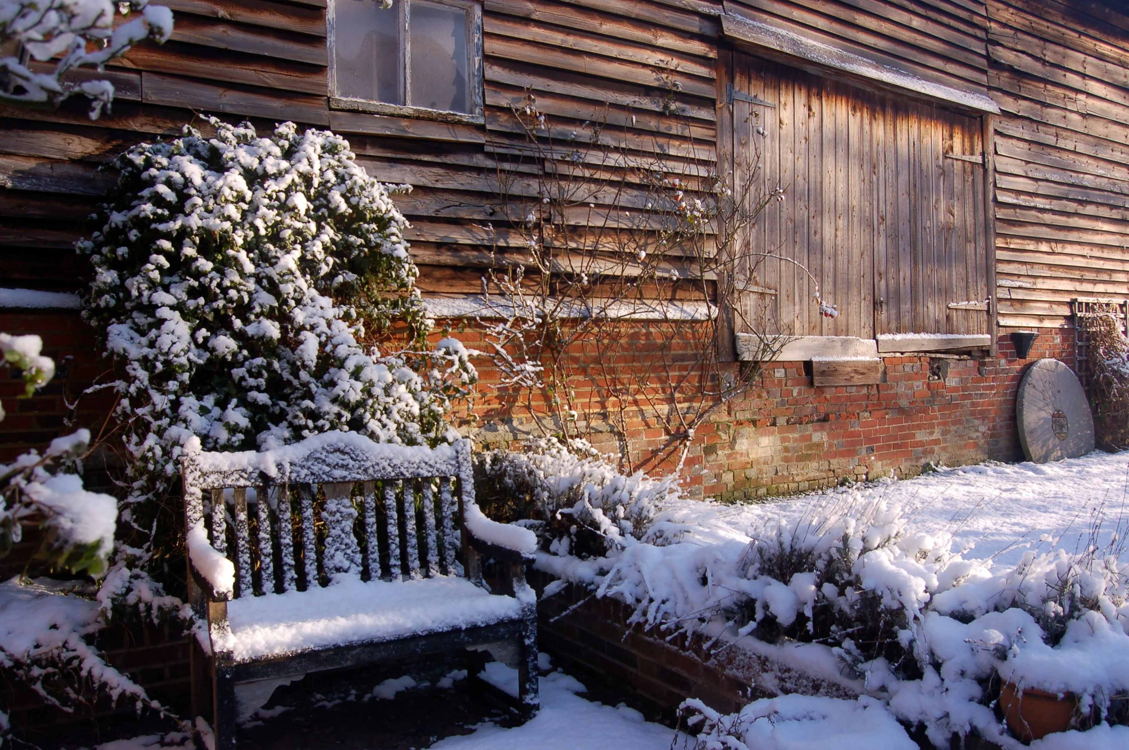 A wooden bench covered in snow sits beside a snow-dusted brick wall next to a weathered wooden barn.
