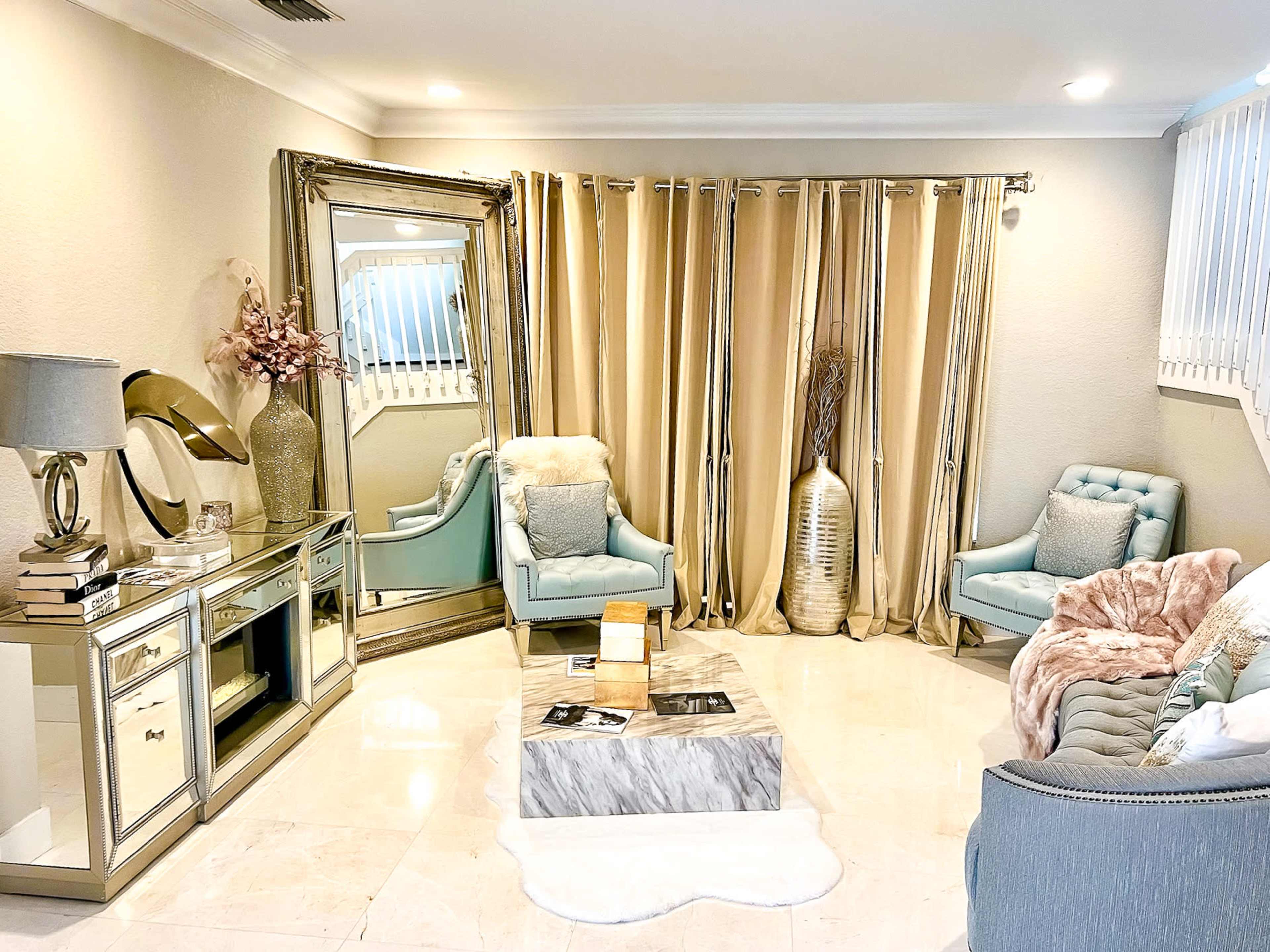 The living room features two light blue chairs, a gray sofa, a decorative mirror, and a marble coffee table, all arranged around a plush white area rug.