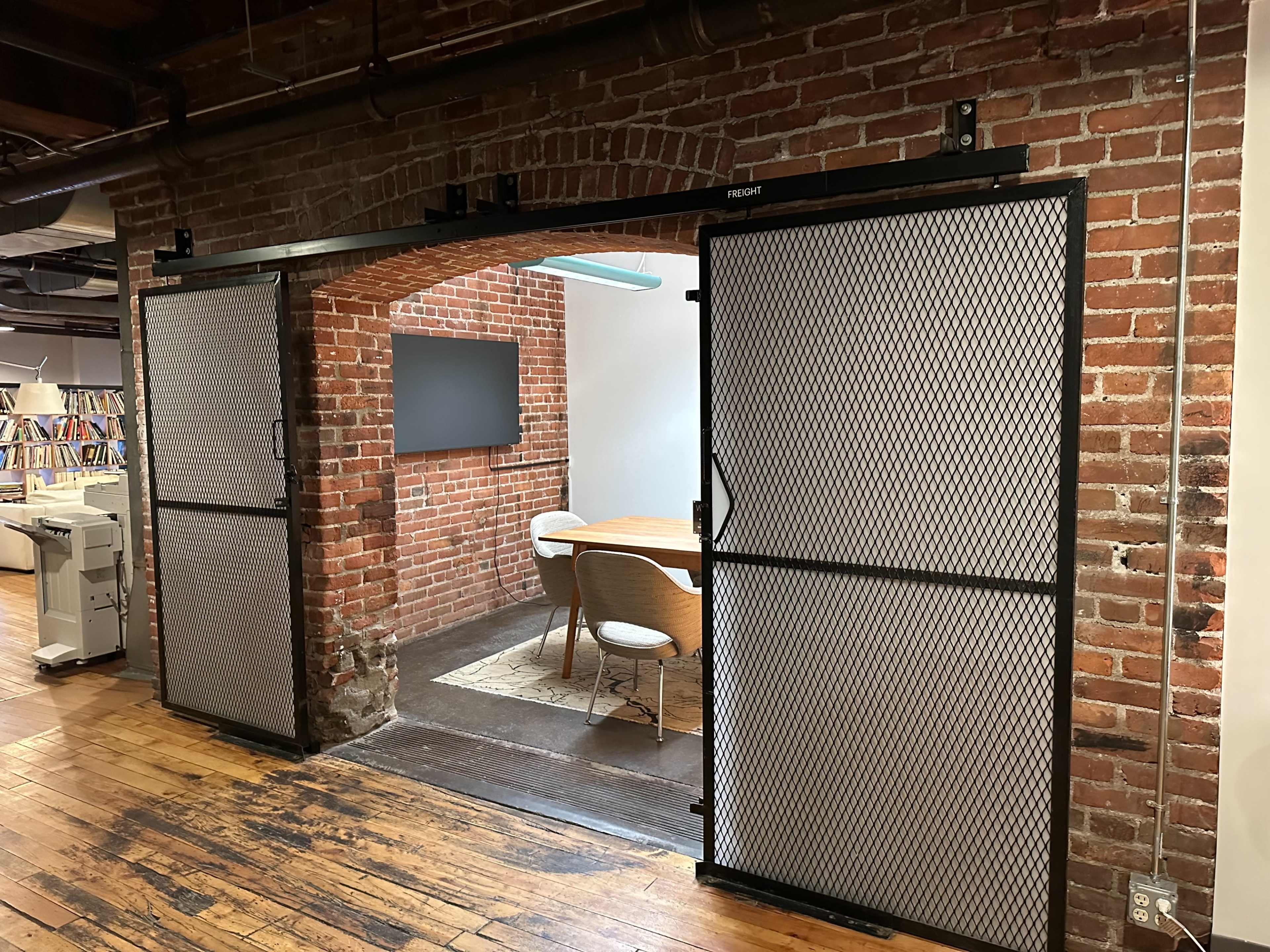 An open entryway with sliding metal mesh doors leading to a meeting room, surrounded by exposed brick walls and wooden flooring.