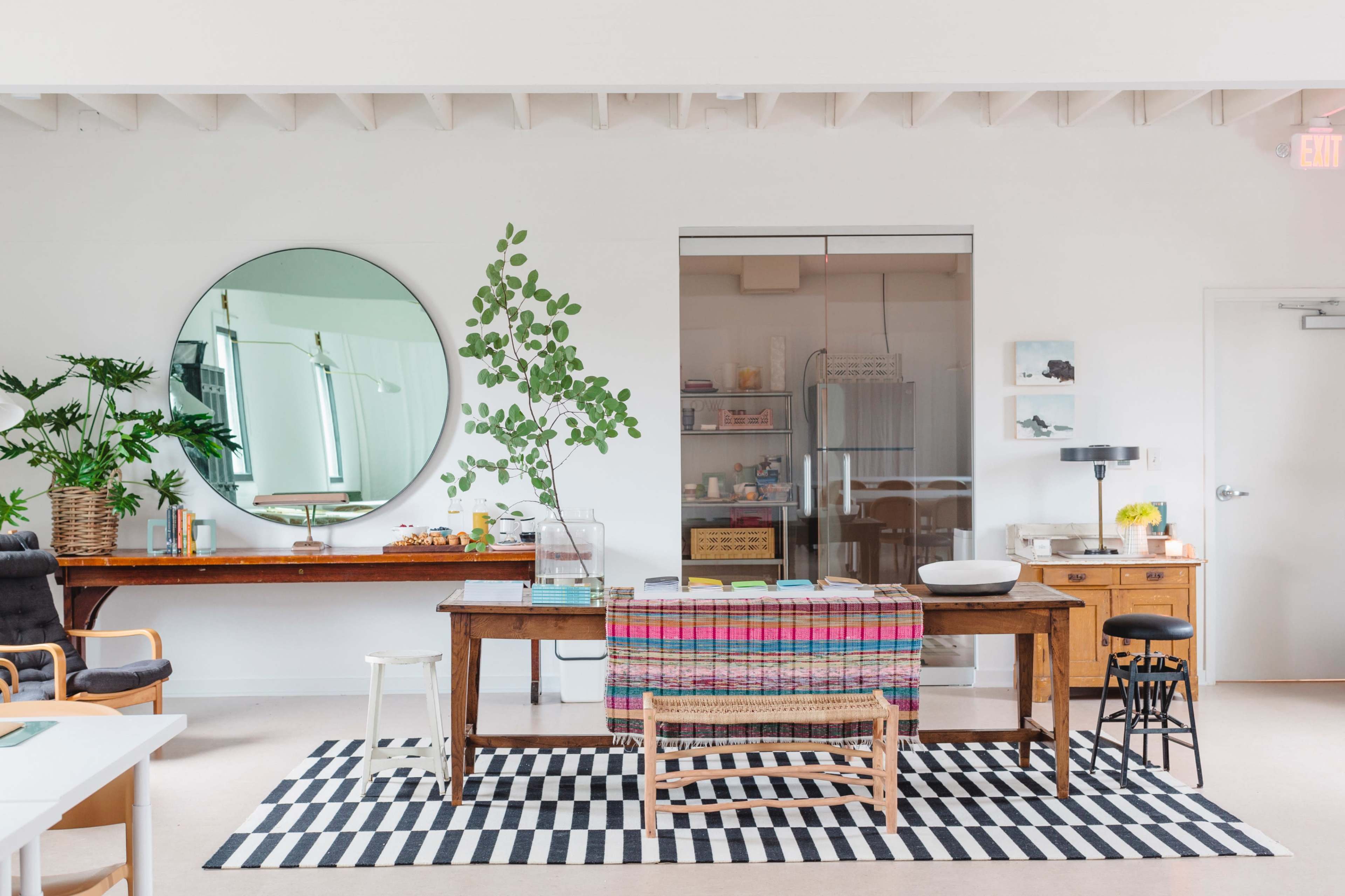 A modern workspace featuring a wooden table draped with a colorful throw, surrounded by various plants, a round mirror, and a glass partition separating a kitchen area.