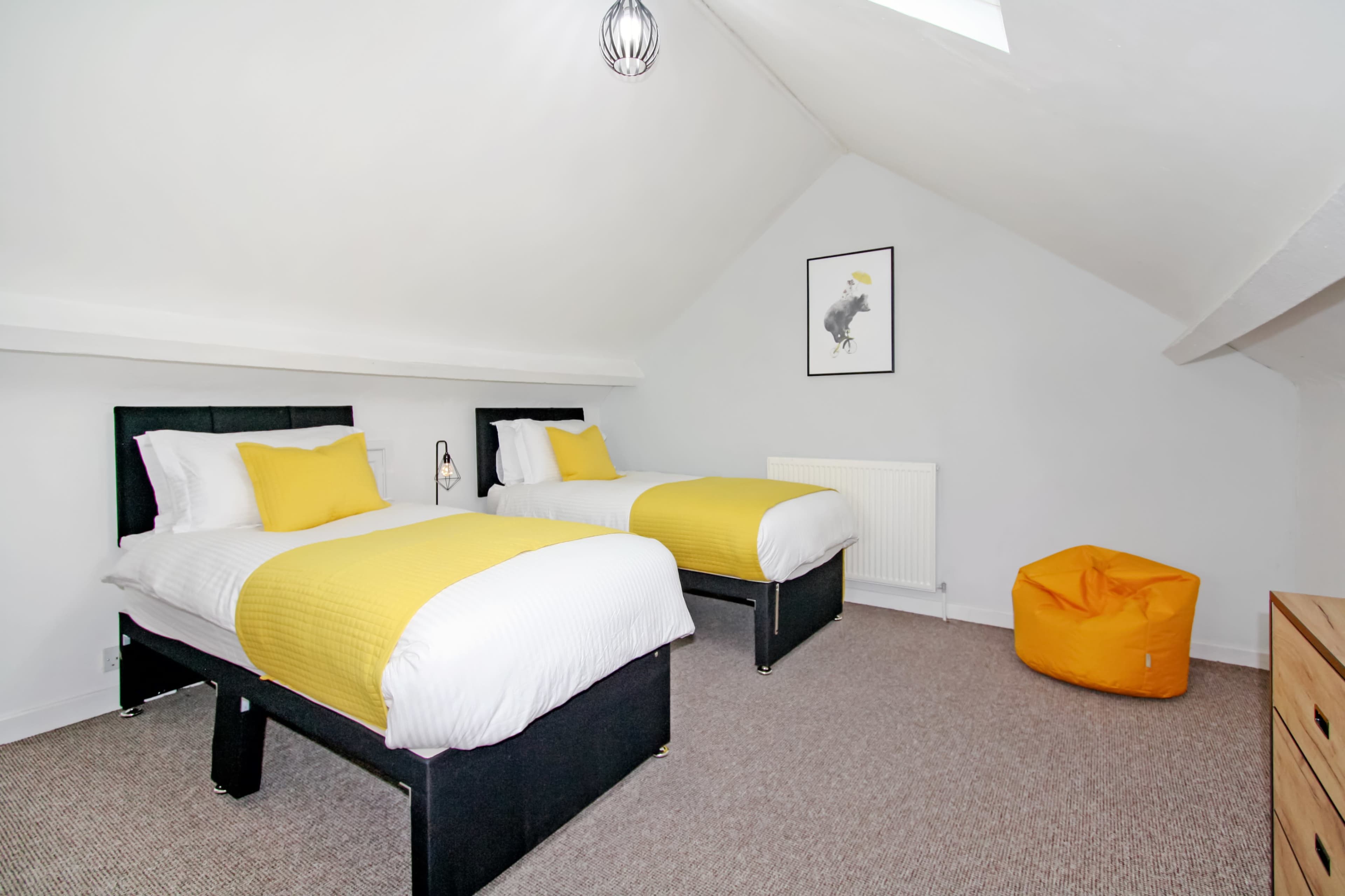 The image shows a room with two twin beds dressed in white linens and yellow accents, a small orange bean bag, and a wall-mounted heater.