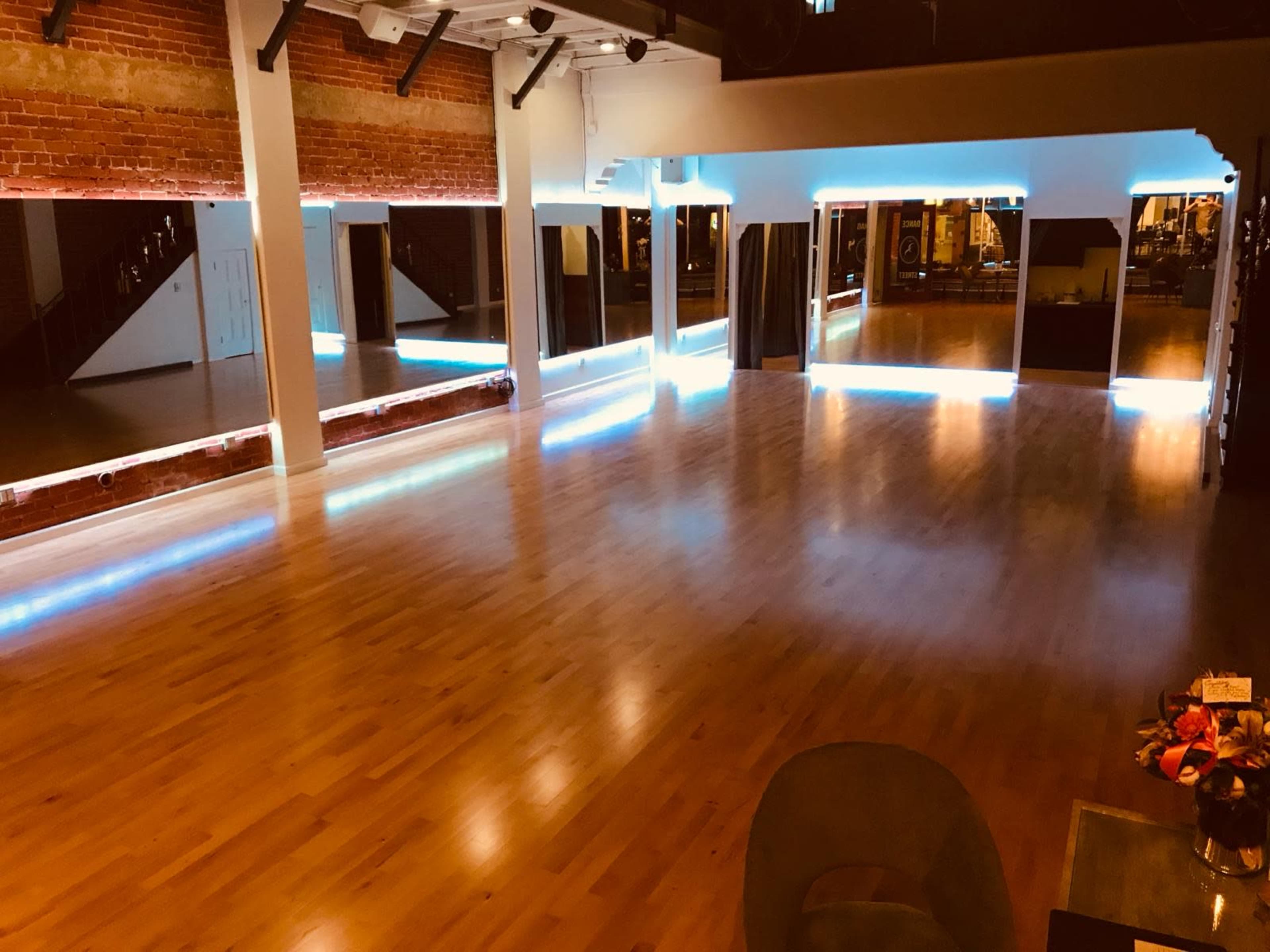 The image shows a spacious dance studio with hardwood floors and illuminated mirrors along the walls.