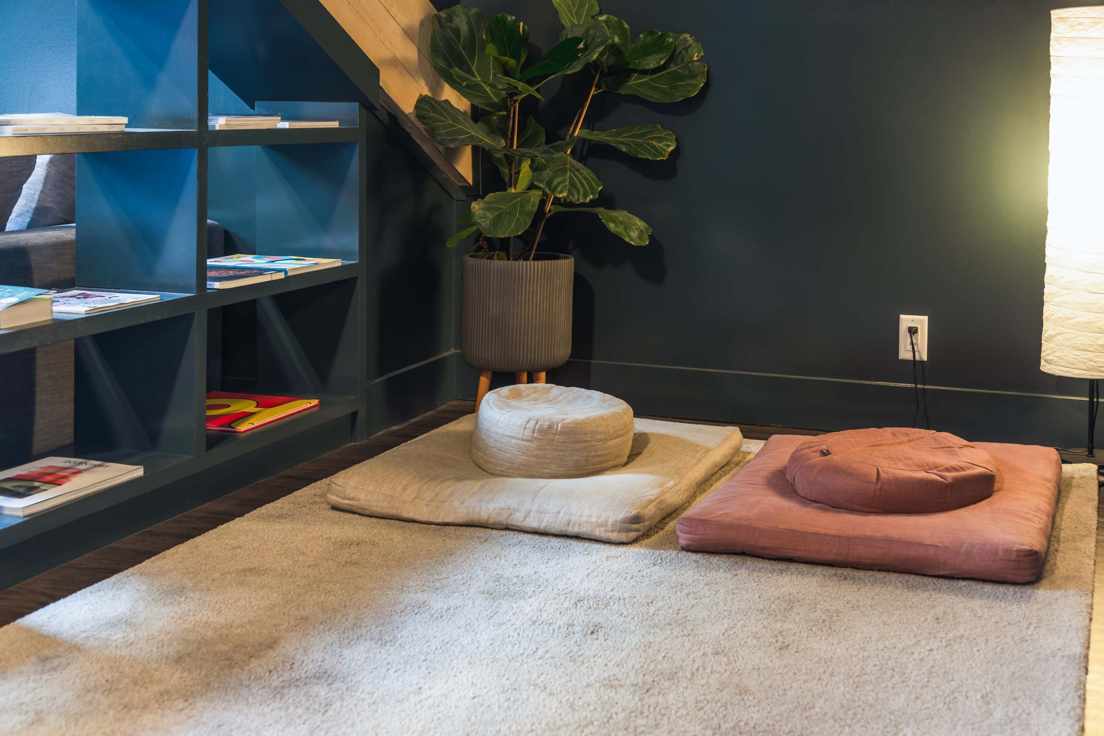 The image shows a cozy corner under a staircase featuring floor cushions, a potted plant, a soft rug, and a lamp beside a shelf with magazines.