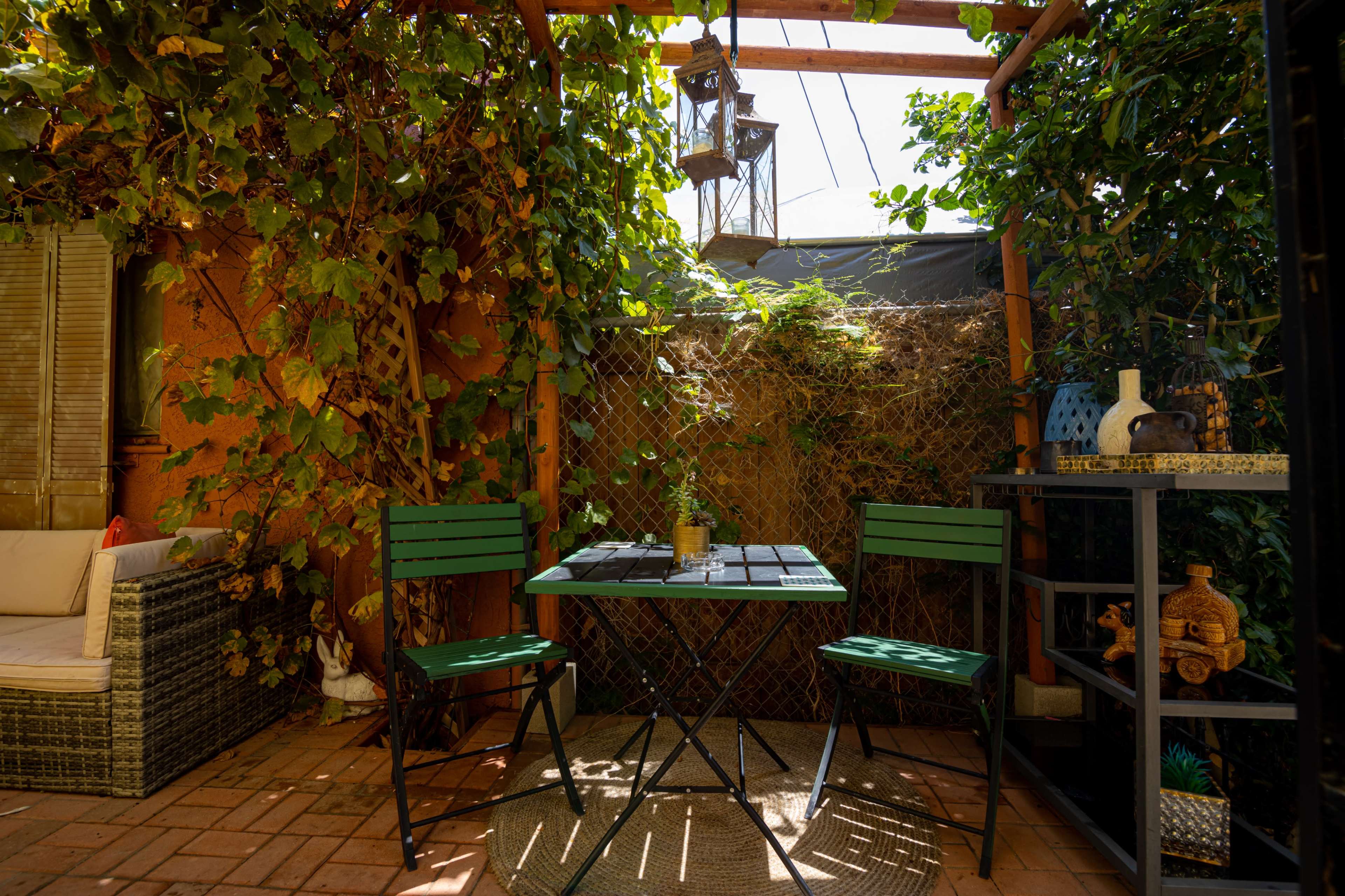 A small outdoor seating area featuring a bistro table and two green chairs, surrounded by lush greenery and decorative elements.