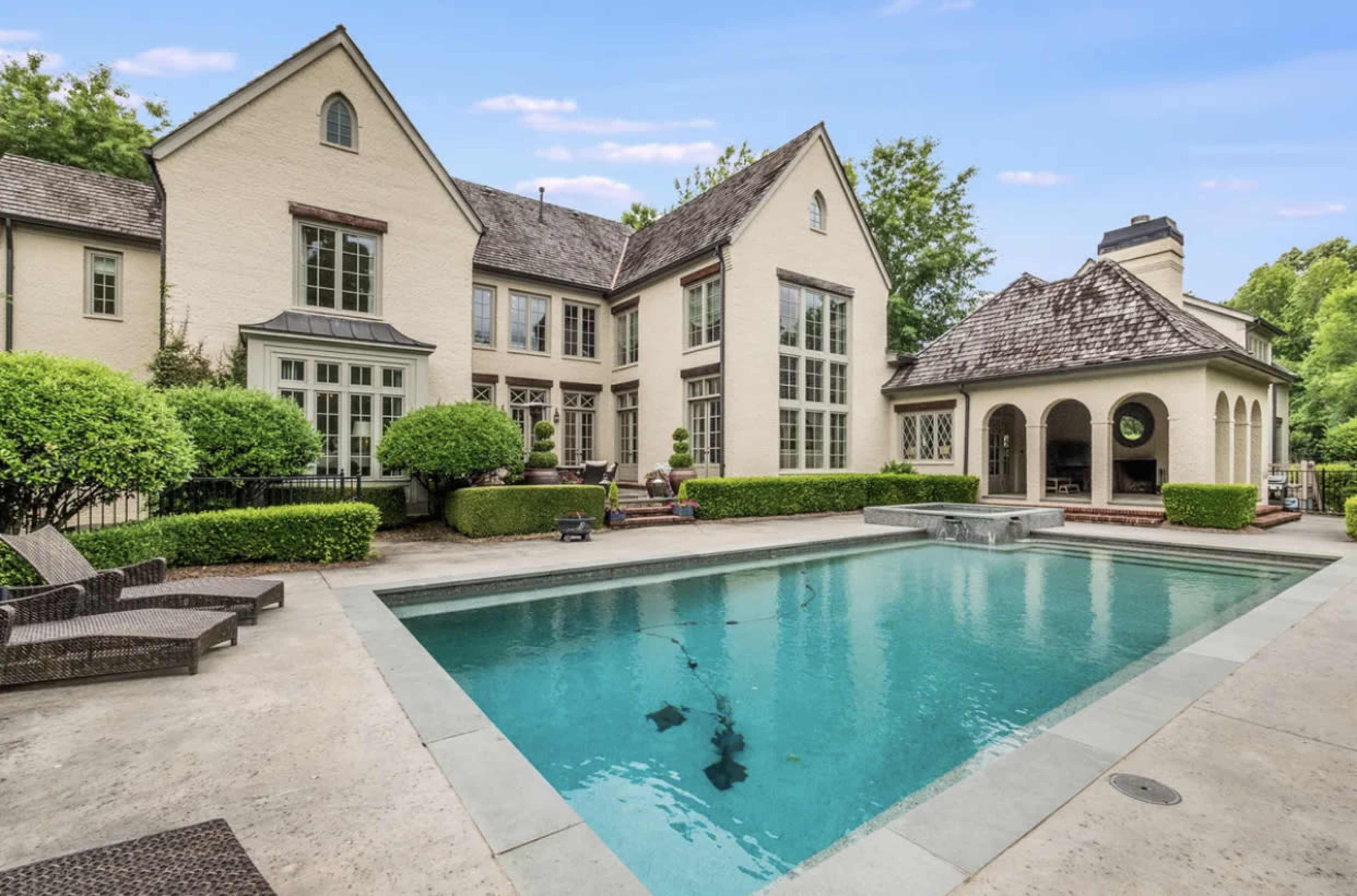 A large house with a pool in the backyard, surrounded by neatly trimmed hedges and a separate structure that appears to be a patio or gazebo.
