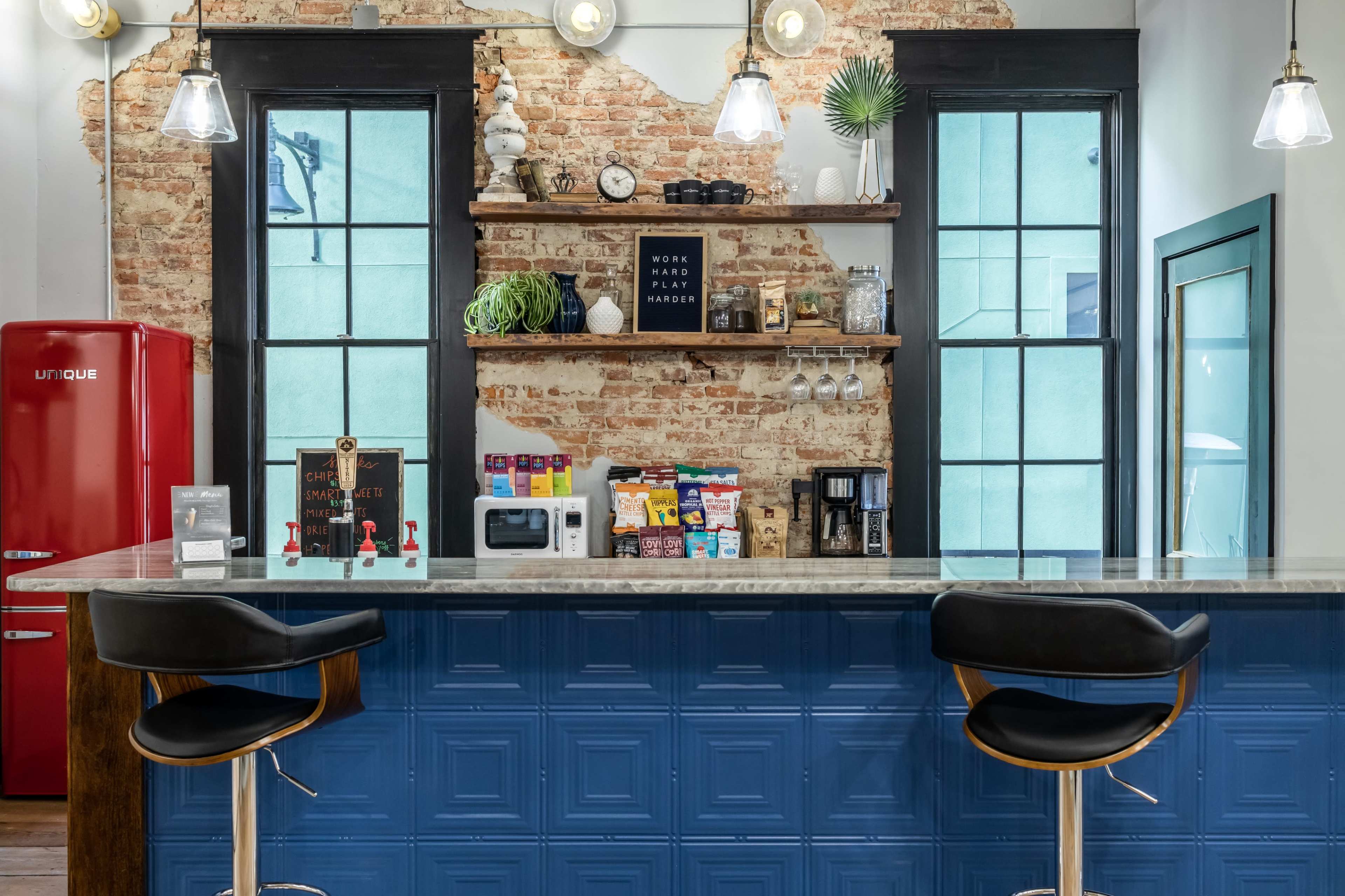 A modern kitchen features a red refrigerator, a granite countertop with bar stools, and shelves displaying various kitchen items against a brick wall.