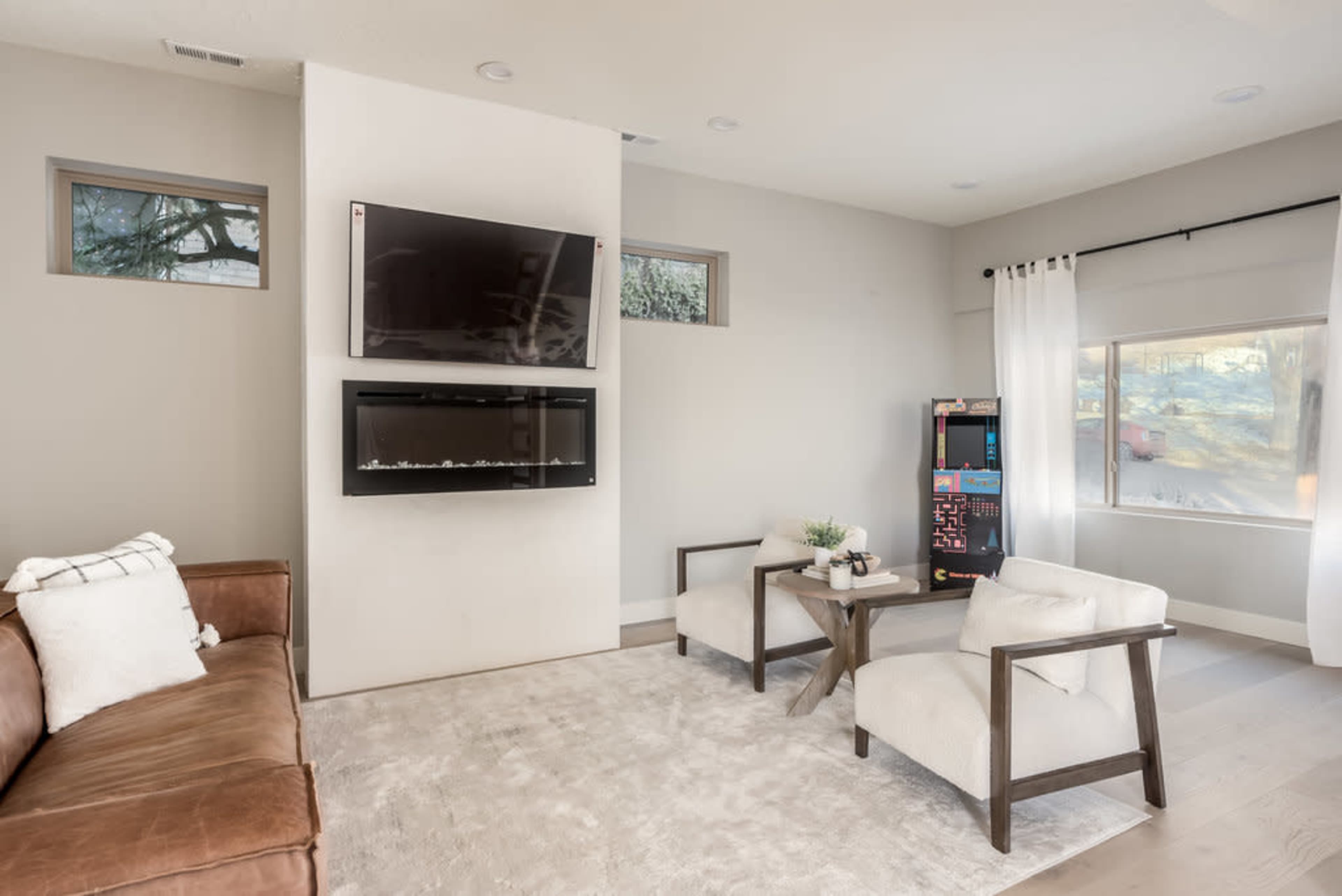 A modern living room with a brown couch, a wall-mounted television, a wall-mounted fireplace, and a small seating area with two chairs beside a large window.
