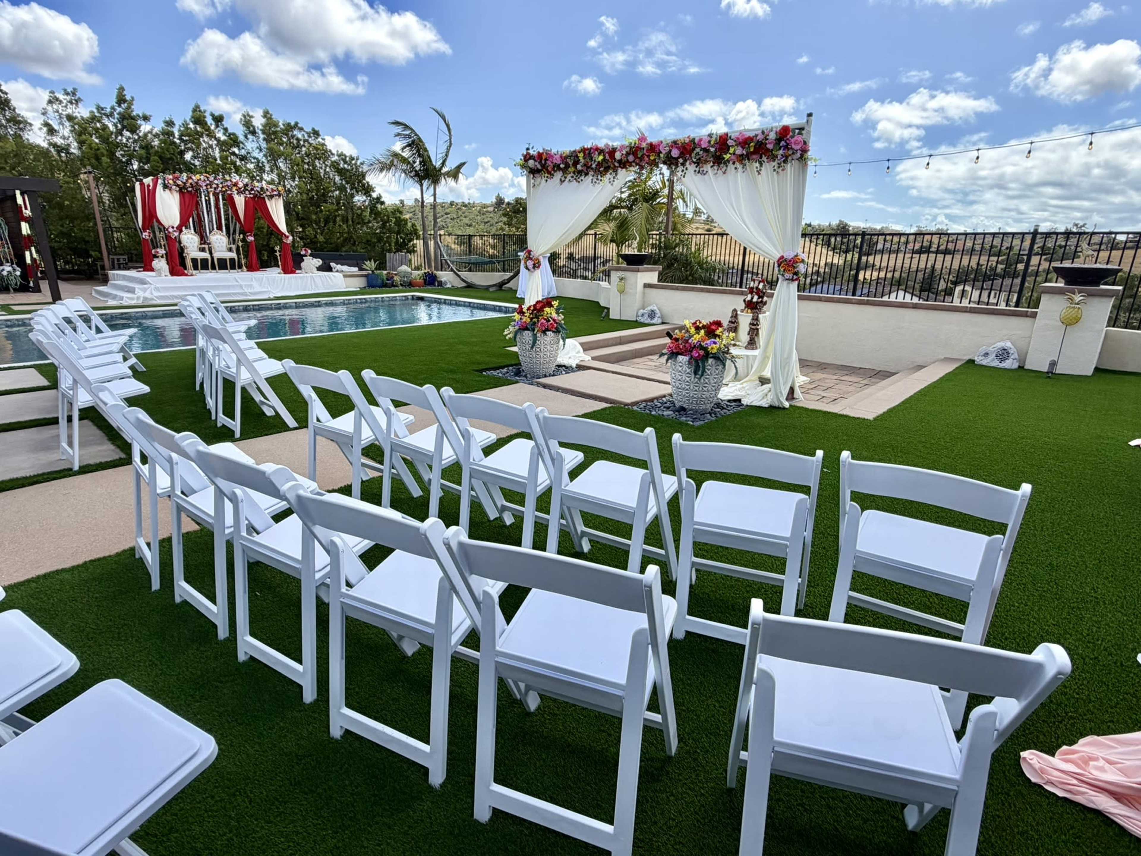 A wedding ceremony setup is displayed outdoors, featuring white chairs arranged in rows facing a decorated altar by a pool, with floral arrangements and a scenic backdrop.