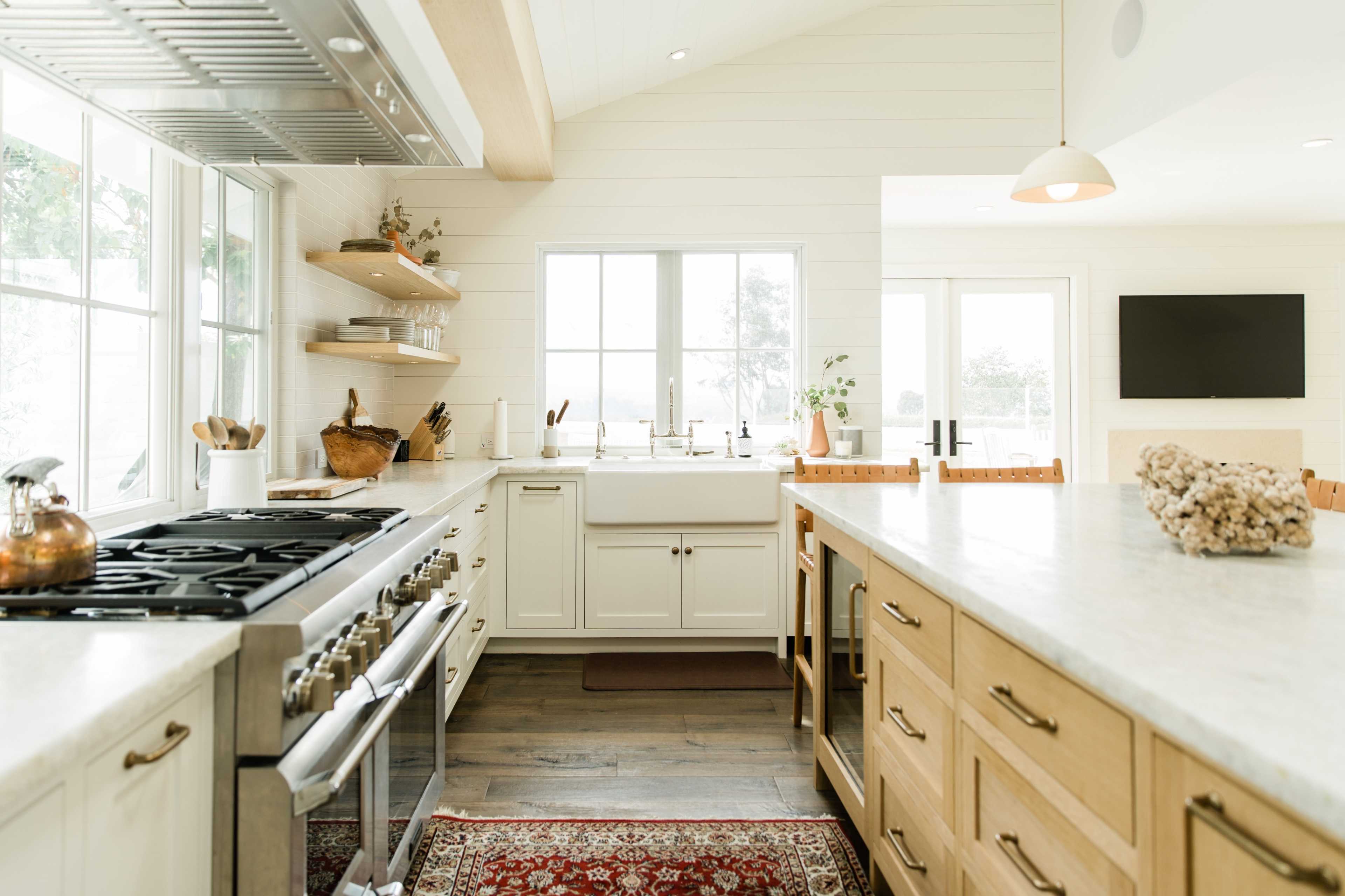 A modern kitchen with a large island, white cabinetry, wooden shelving, and large windows illuminating the space.