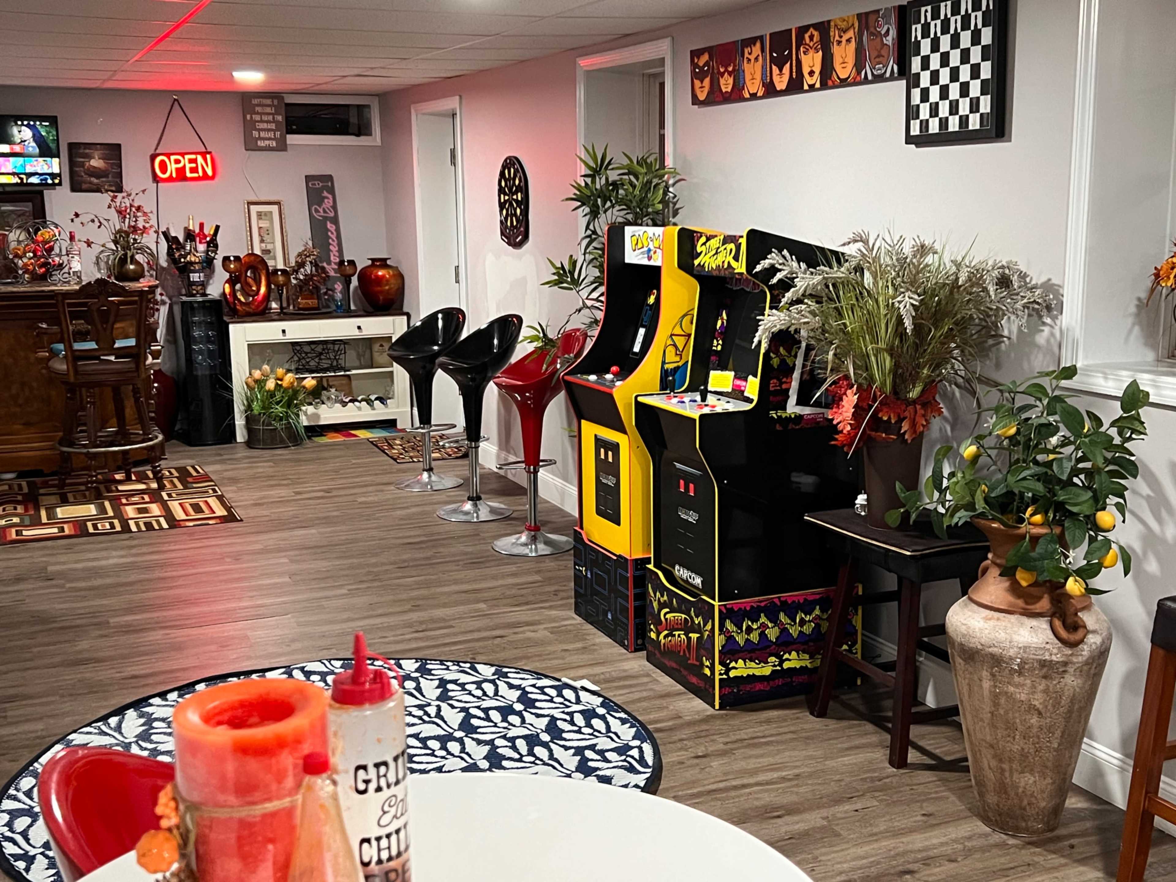 The scene features a cozy basement game room with vintage arcade machines, a bar area, and festive autumn decorations.