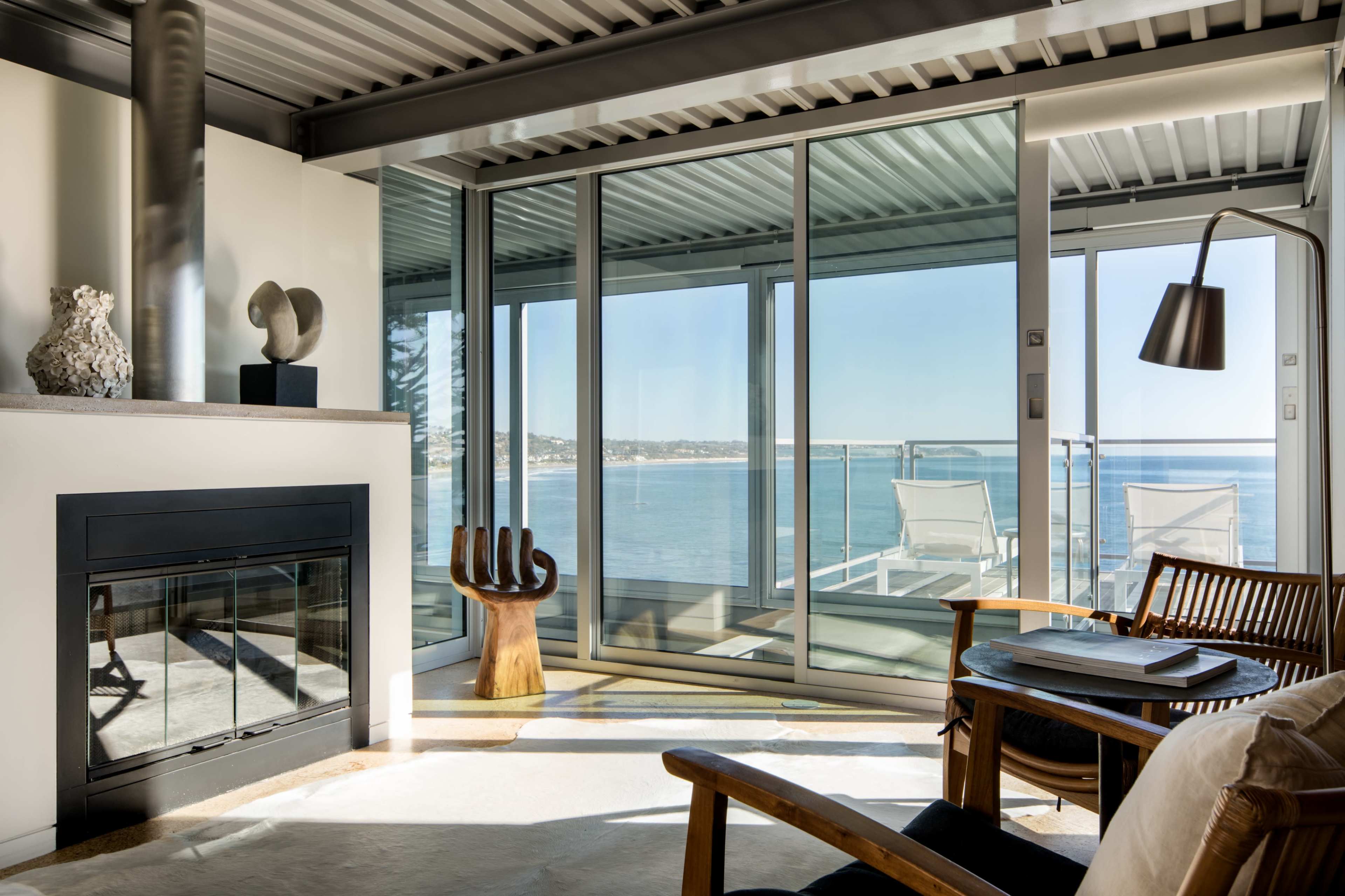 A modern living space with large glass windows overlooking a body of water, featuring a fireplace and wooden furniture.