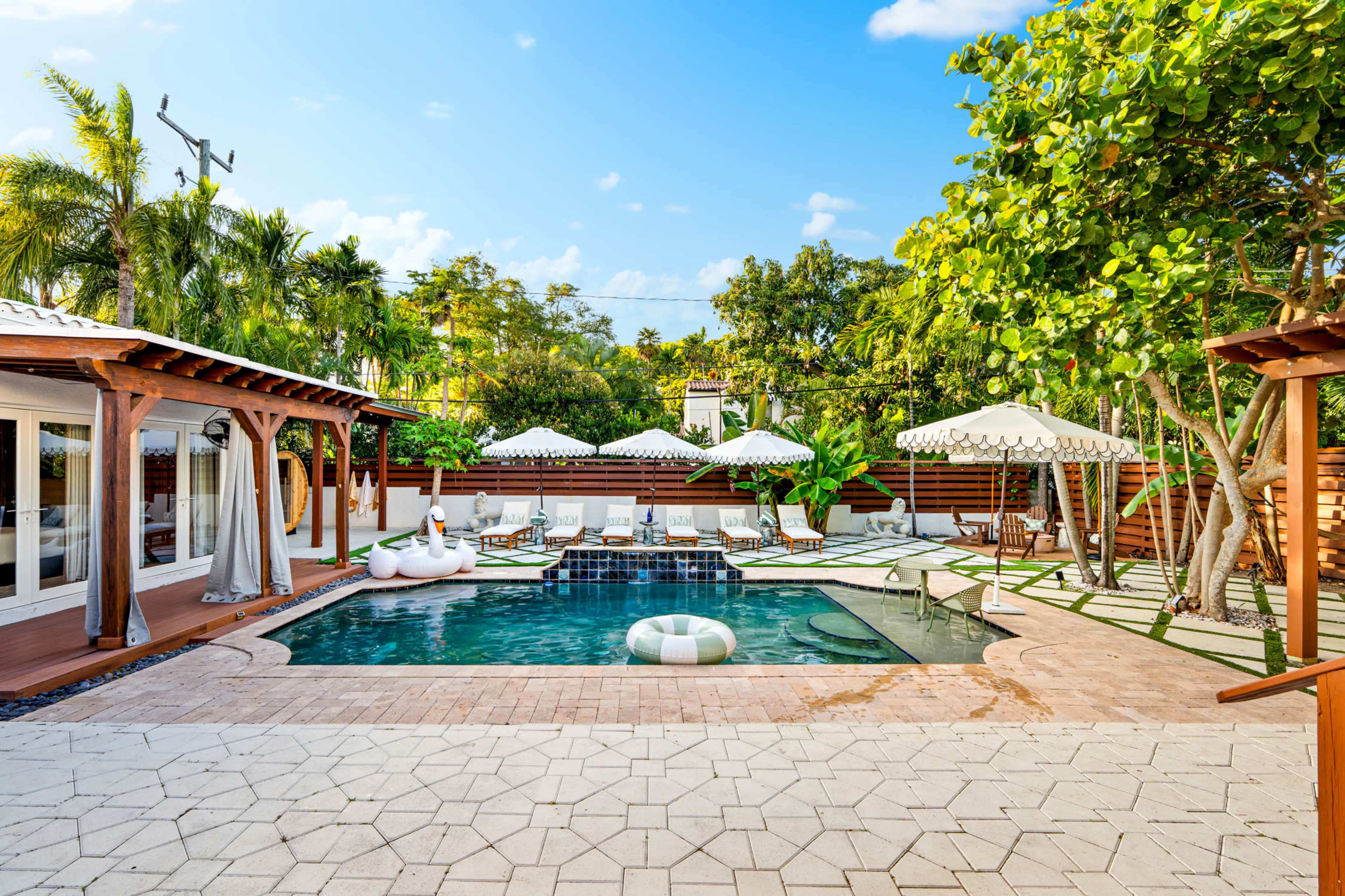 The image shows a spacious outdoor pool area surrounded by sun loungers, umbrellas, and tropical greenery.