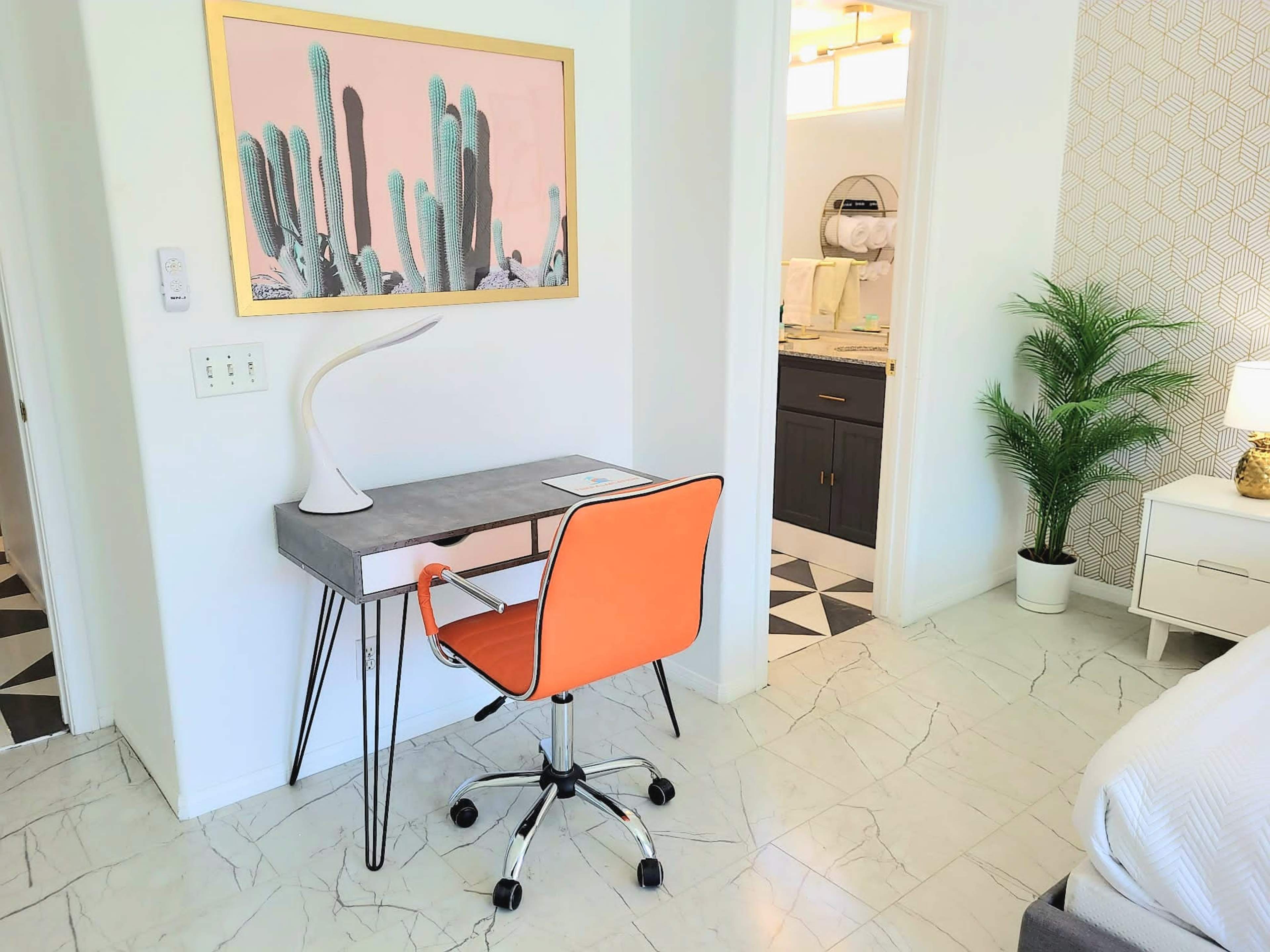 The image shows a modern workspace featuring a gray desk with an orange chair, a cactus-themed wall art, and a view into a bathroom through an open doorway.