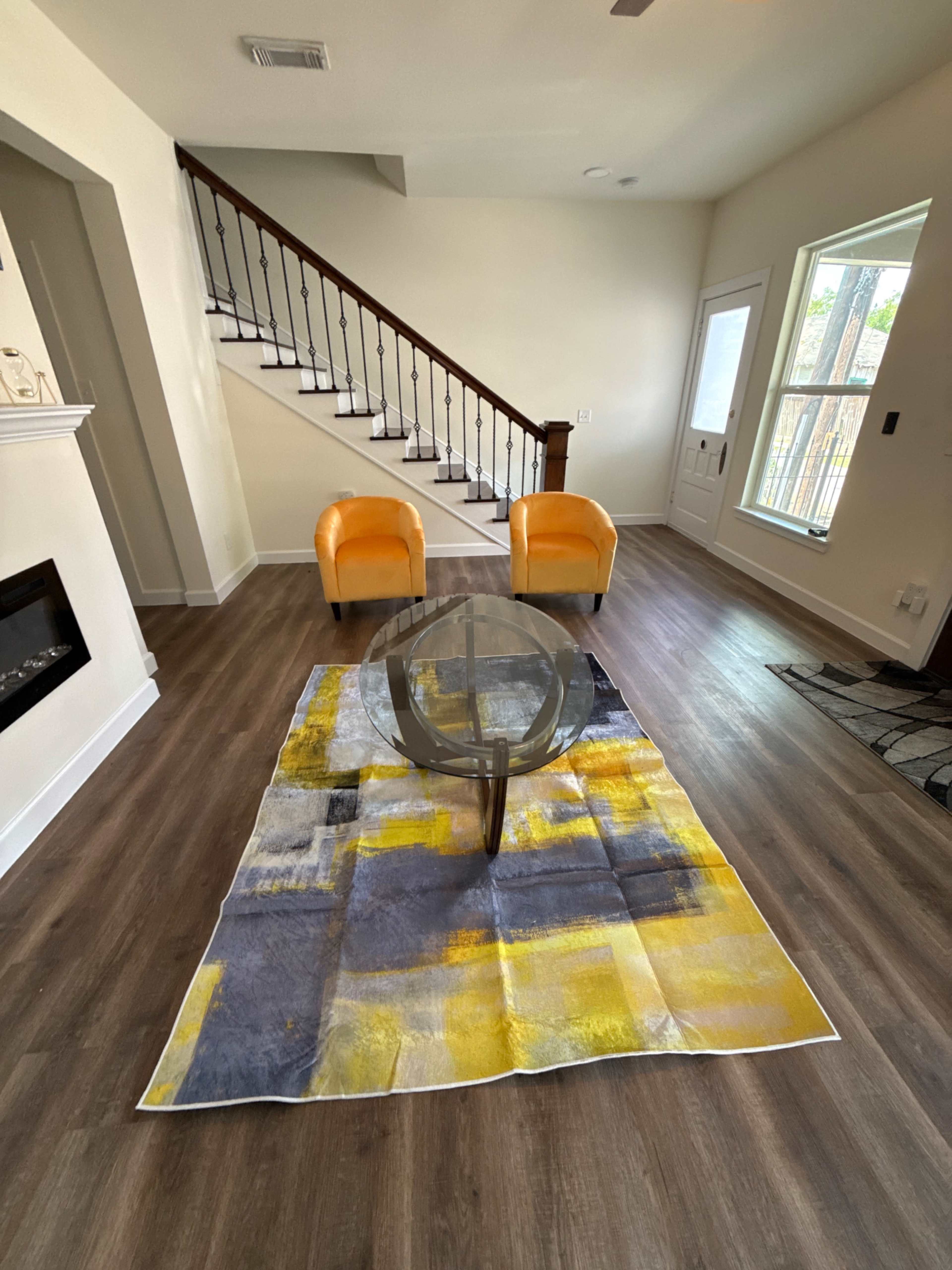 The image shows a modern living room featuring a glass coffee table on a colorful area rug, with two orange chairs and a staircase in the background.