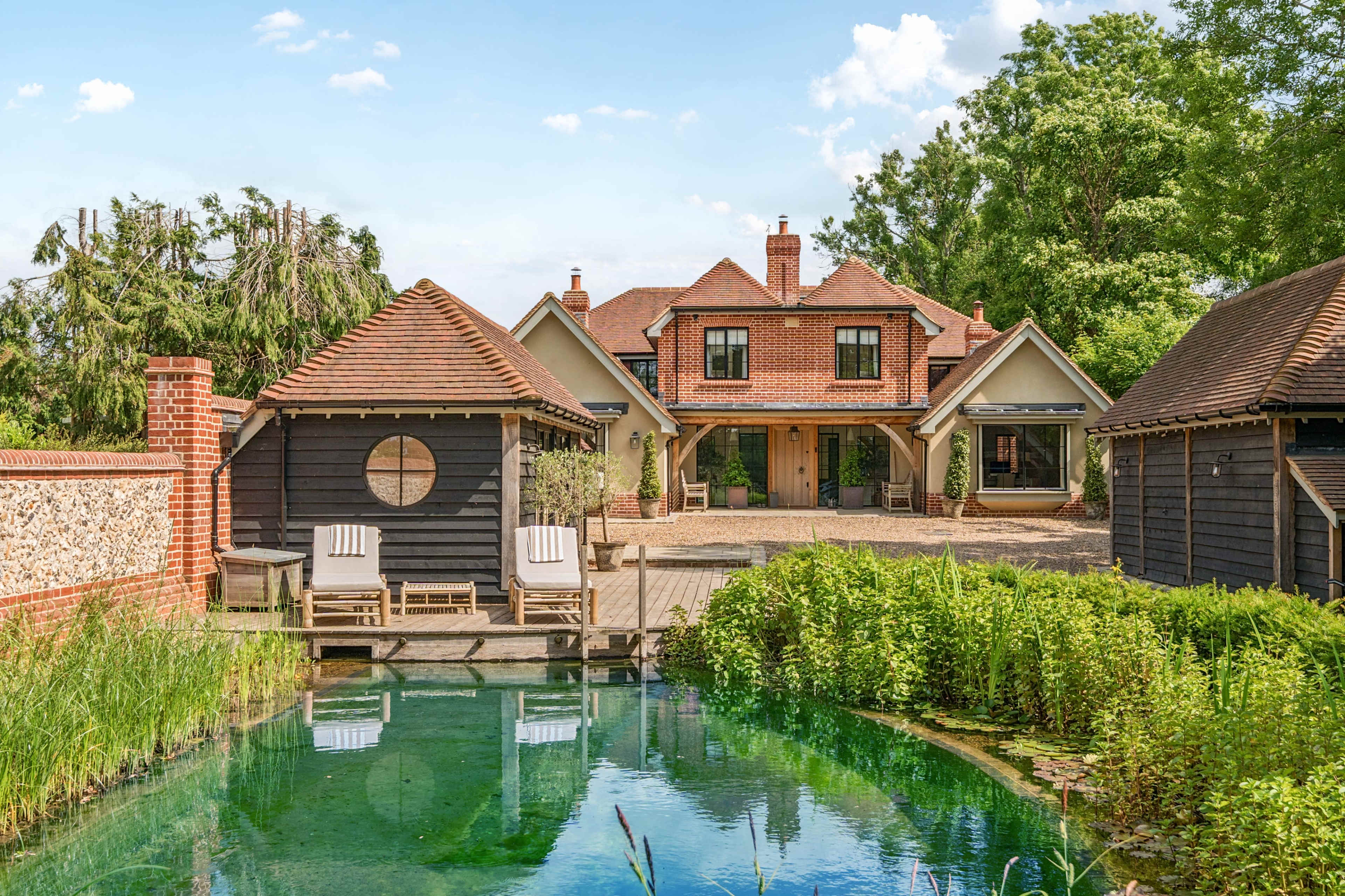 The image shows a house with a central entrance surrounded by several outbuildings, a pond in the foreground, and greenery in the background.