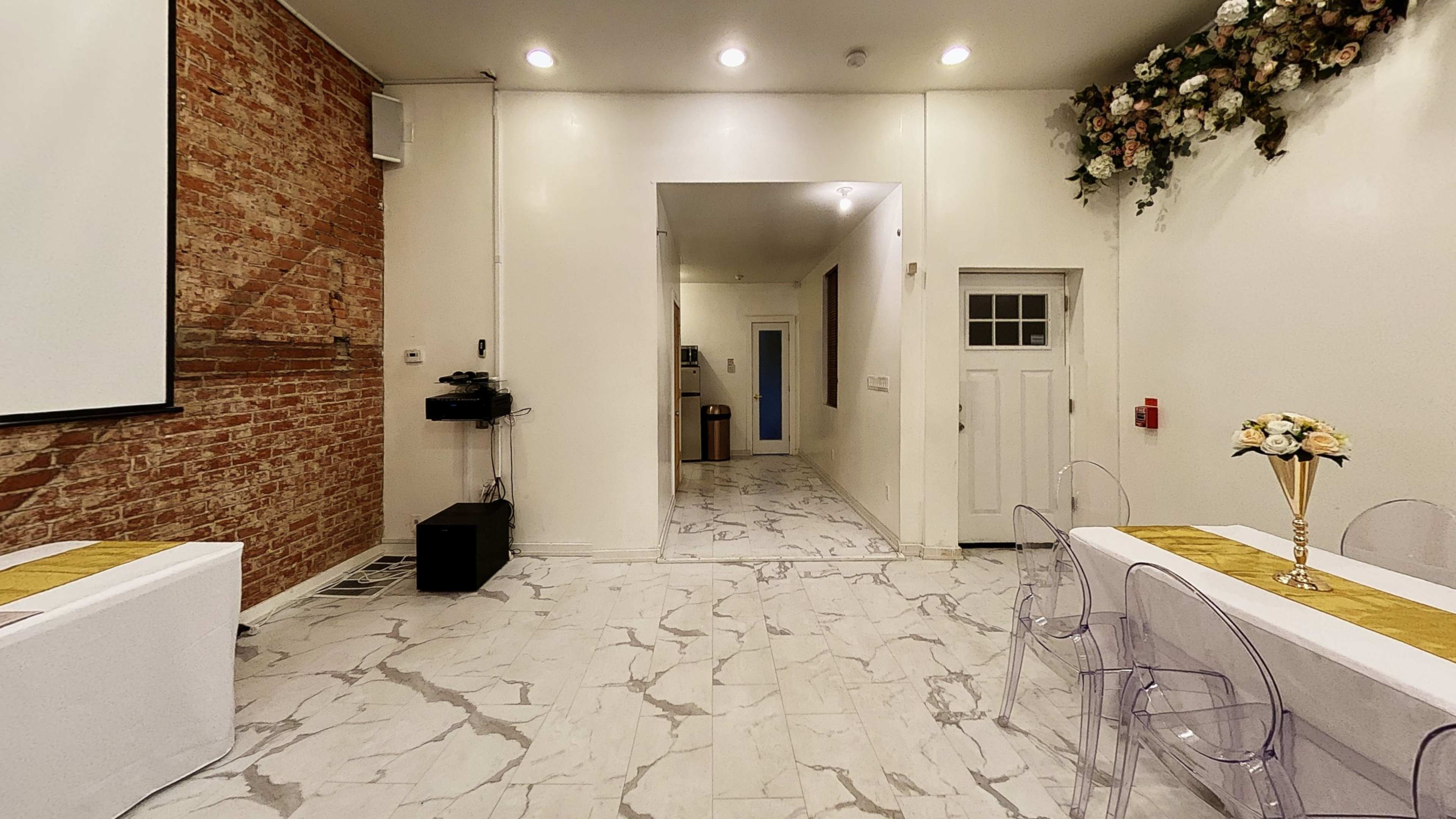 A spacious room with white walls, marble flooring, and a decorative floral arrangement on one side, furnished with transparent chairs and tables set with gold runners.
