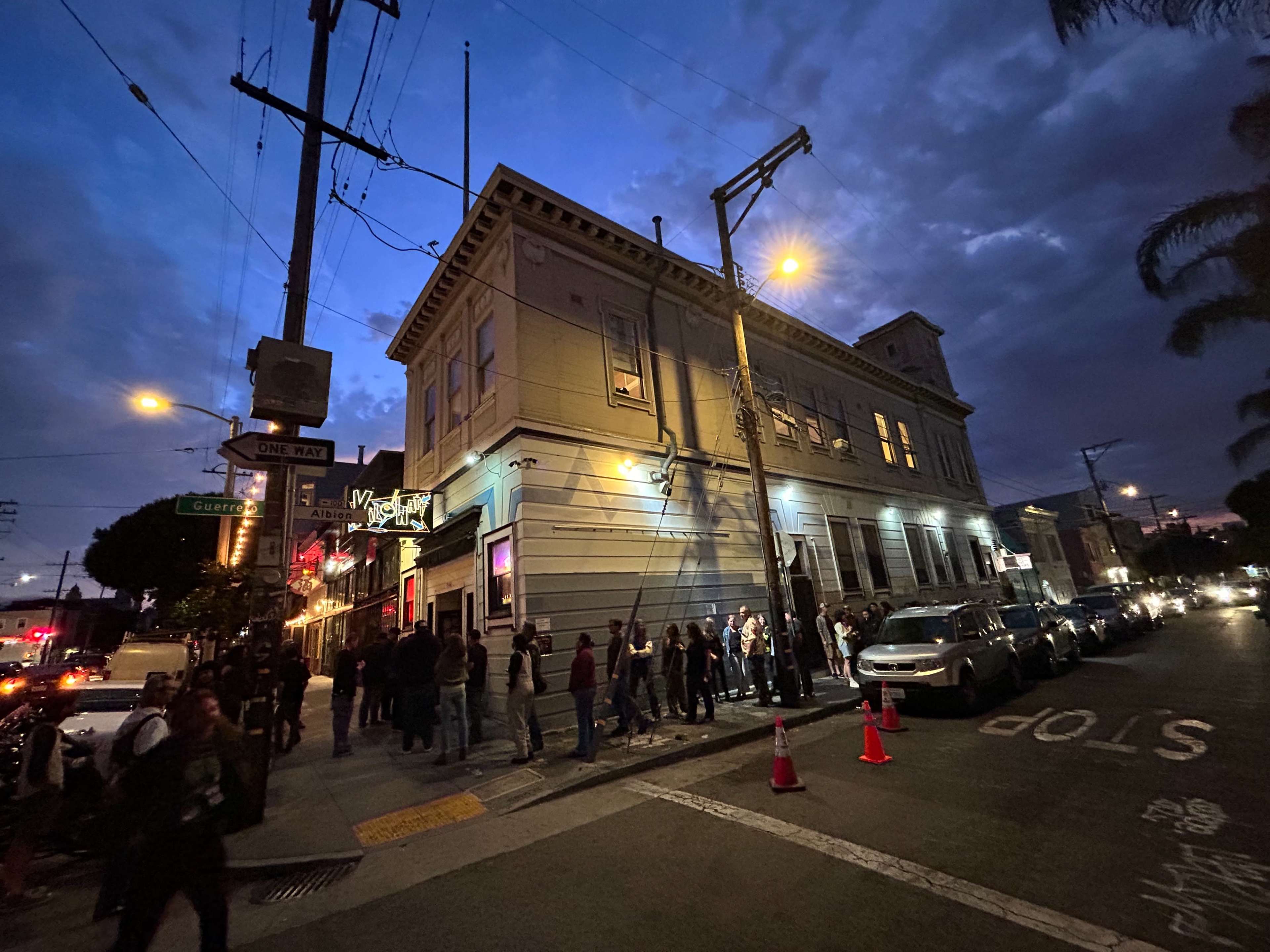 A queue of people stands outside a large building with illuminated windows during dusk on a city street.
