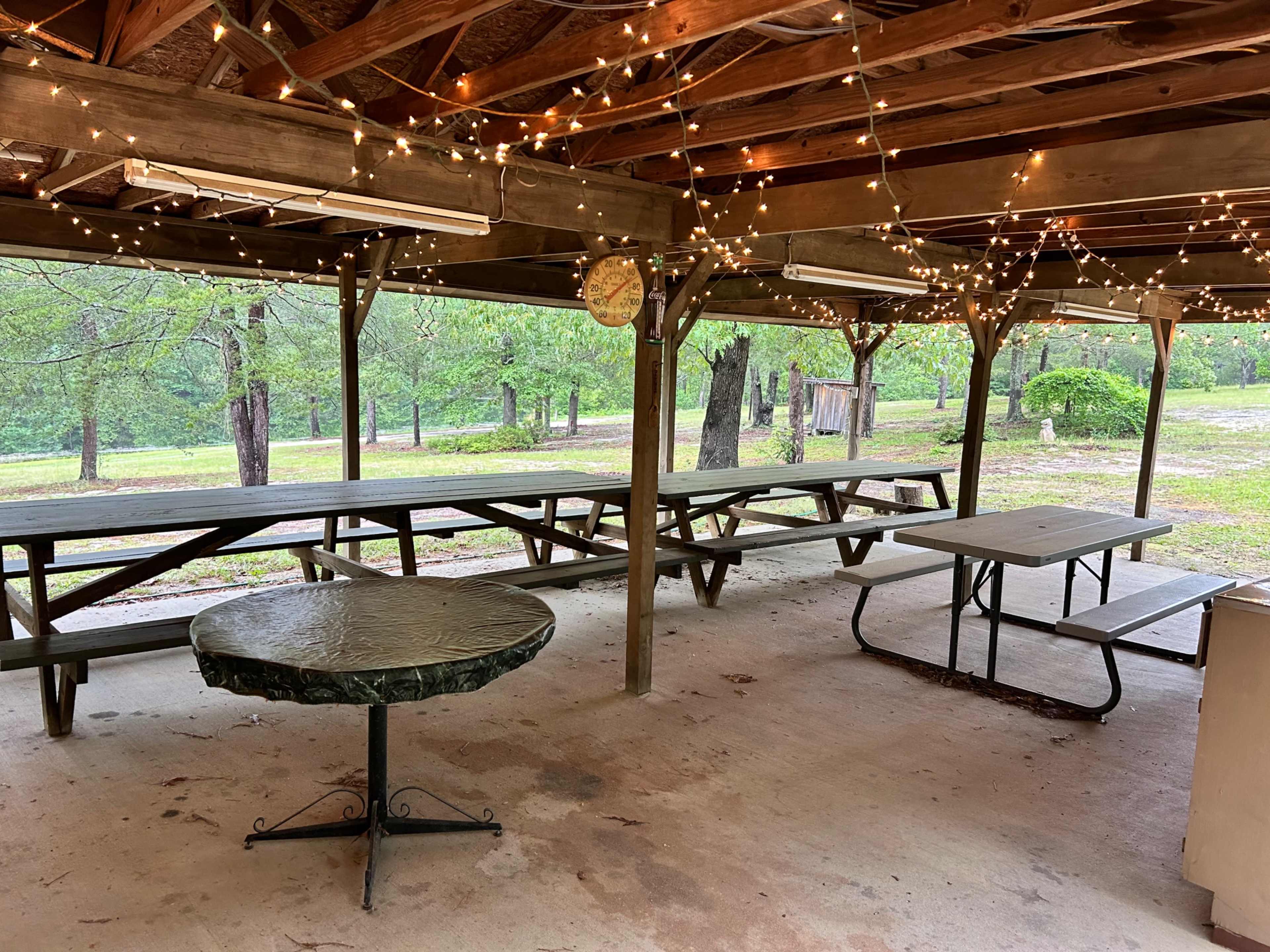 The image shows an open pavilion with wooden beams and string lights, featuring several picnic tables and a round table underneath.