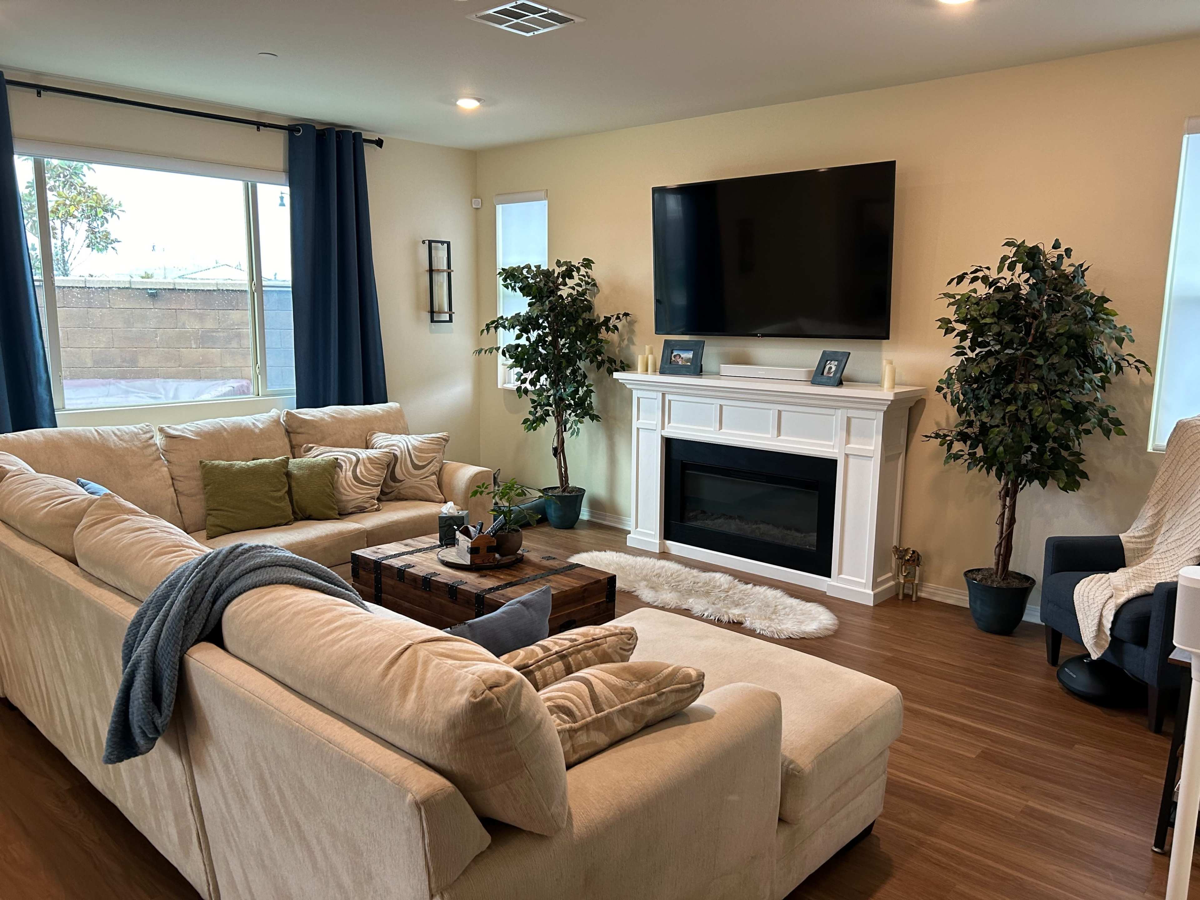 The living room features a large sectional sofa, a coffee table, a television mounted on the wall, and two artificial plants beside a faux fireplace.