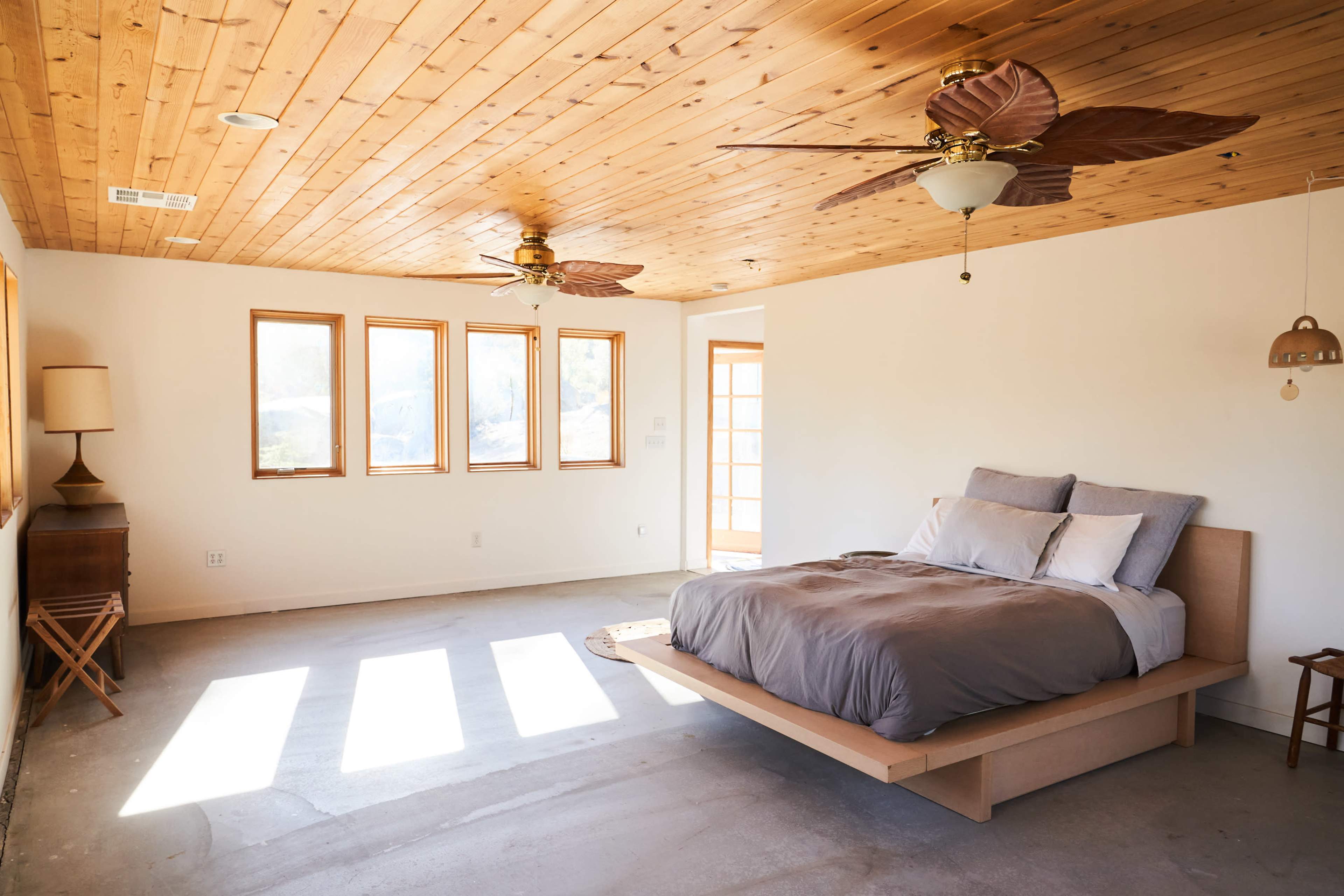 The bedroom features a wooden ceiling, large windows allowing natural light, and a minimalist bed with a gray comforter.