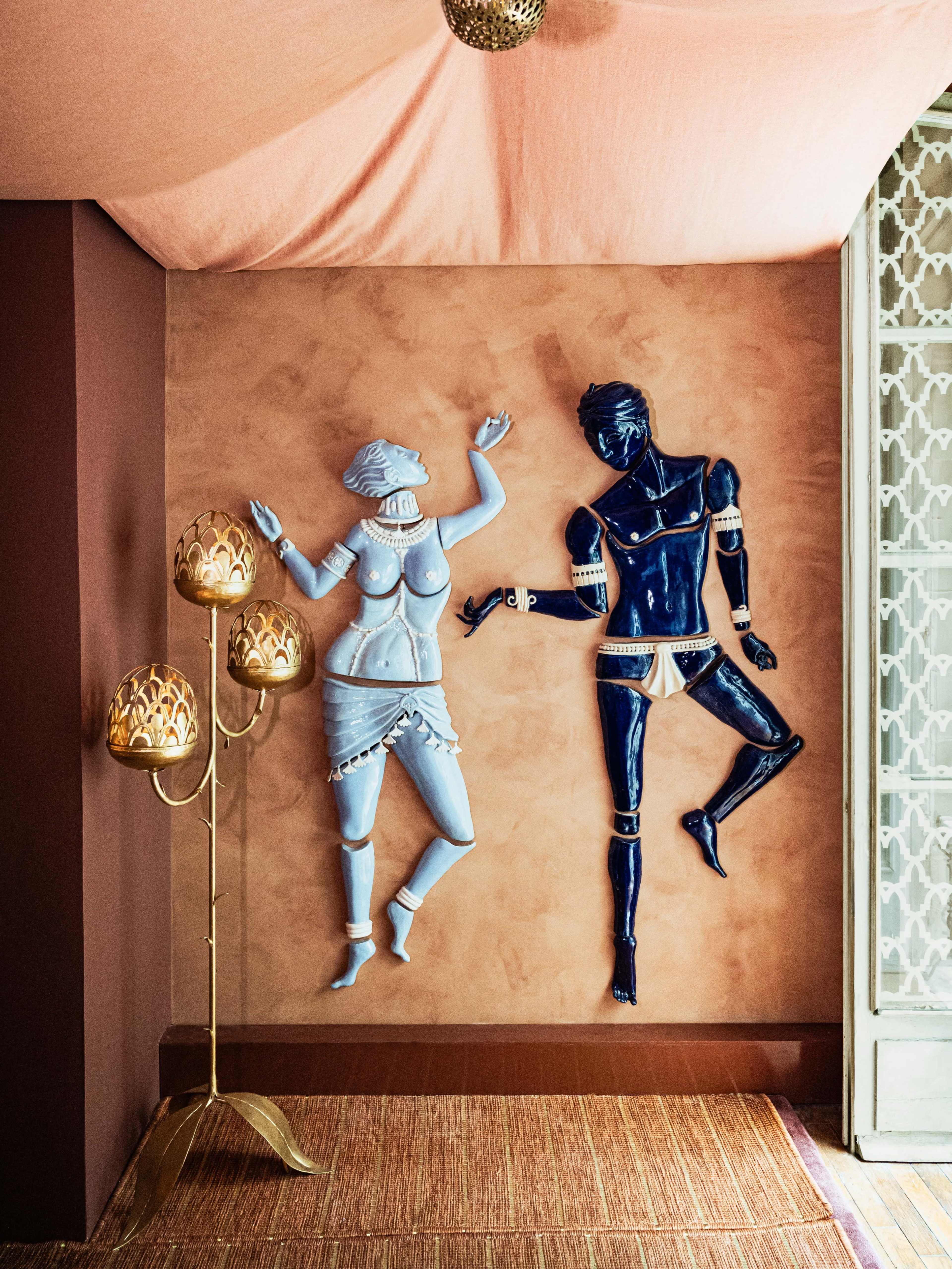 The image shows two large wall sculptures of a couple, one in blue and the other in light blue, set against a textured tan wall, with a floor lamp featuring gold shades nearby.