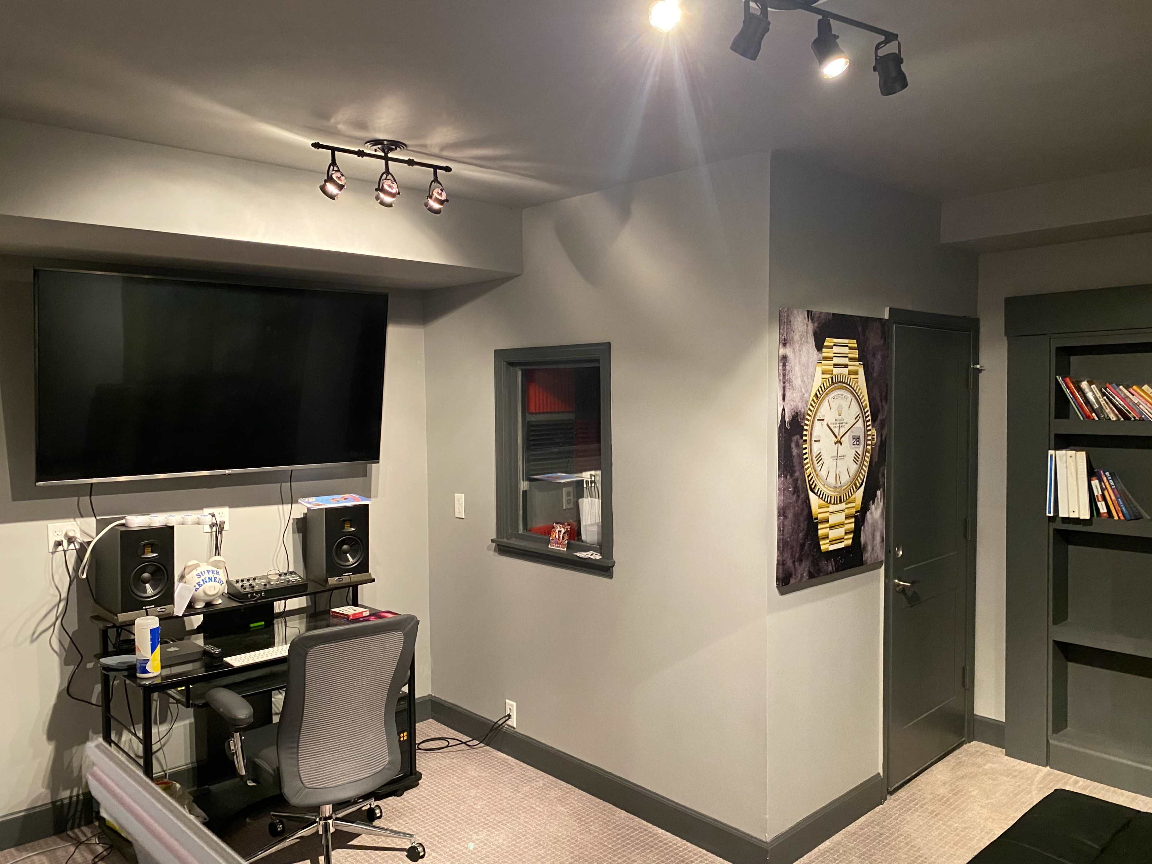 A modern room featuring a desk with speakers, a large wall-mounted TV, a framed clock artwork, and a bookshelf.