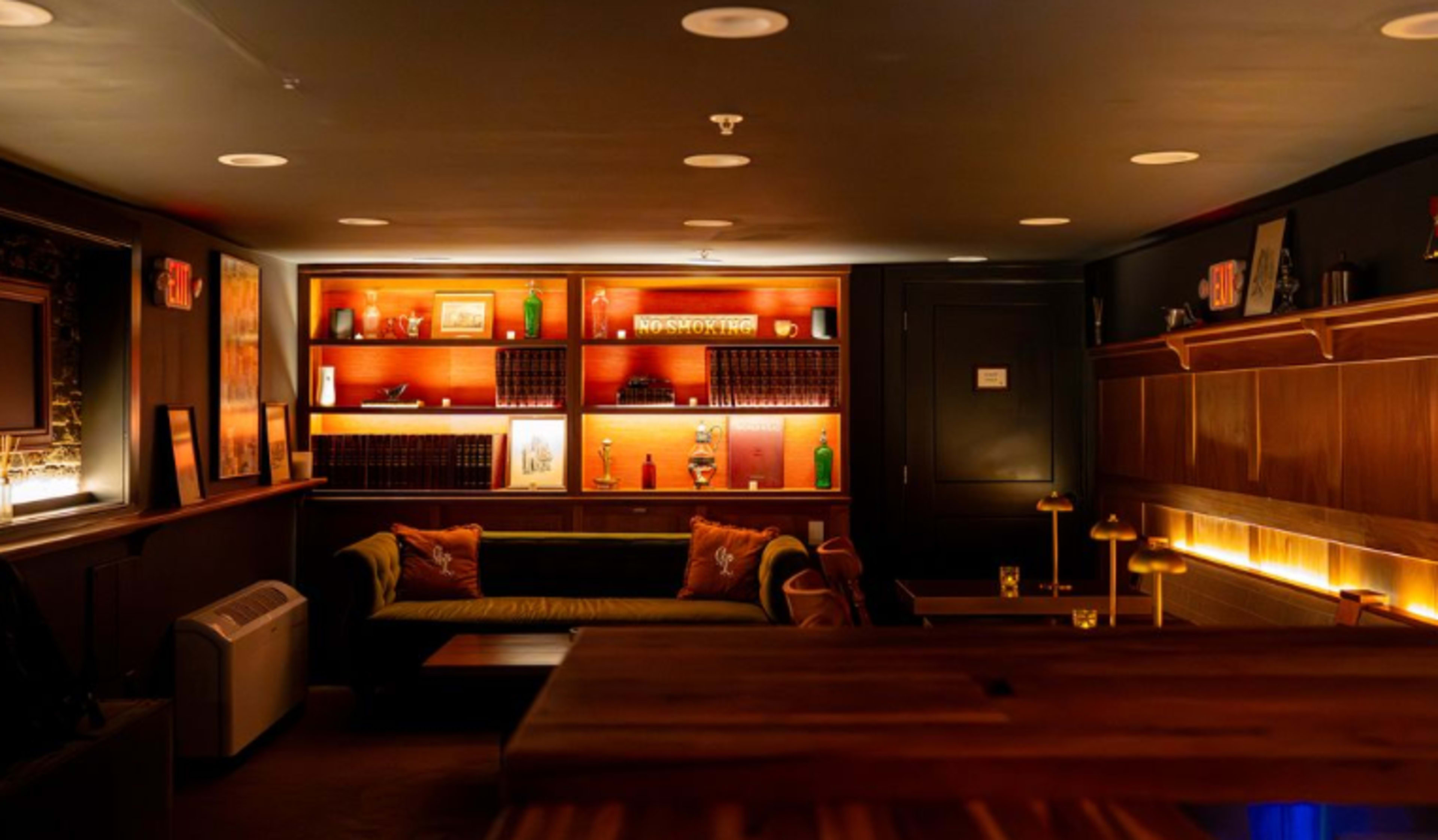 The image shows a dimly lit lounge area featuring a decorative bookshelf, a plush sofa, and a polished wooden bar.