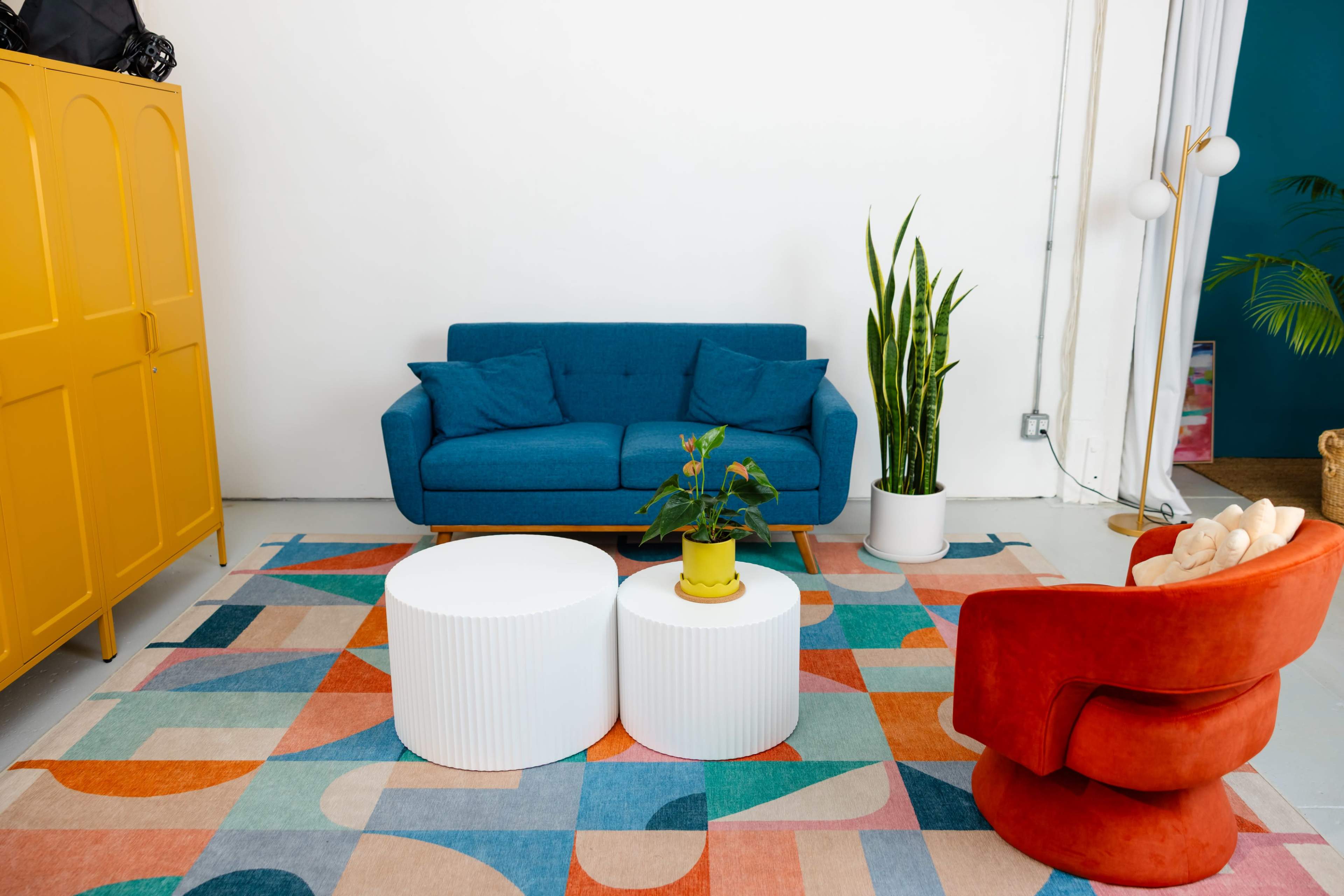 The image shows a modern living room with a blue sofa, two white side tables, a potted plant, and an orange armchair, all set on a colorful geometric rug against a white wall.