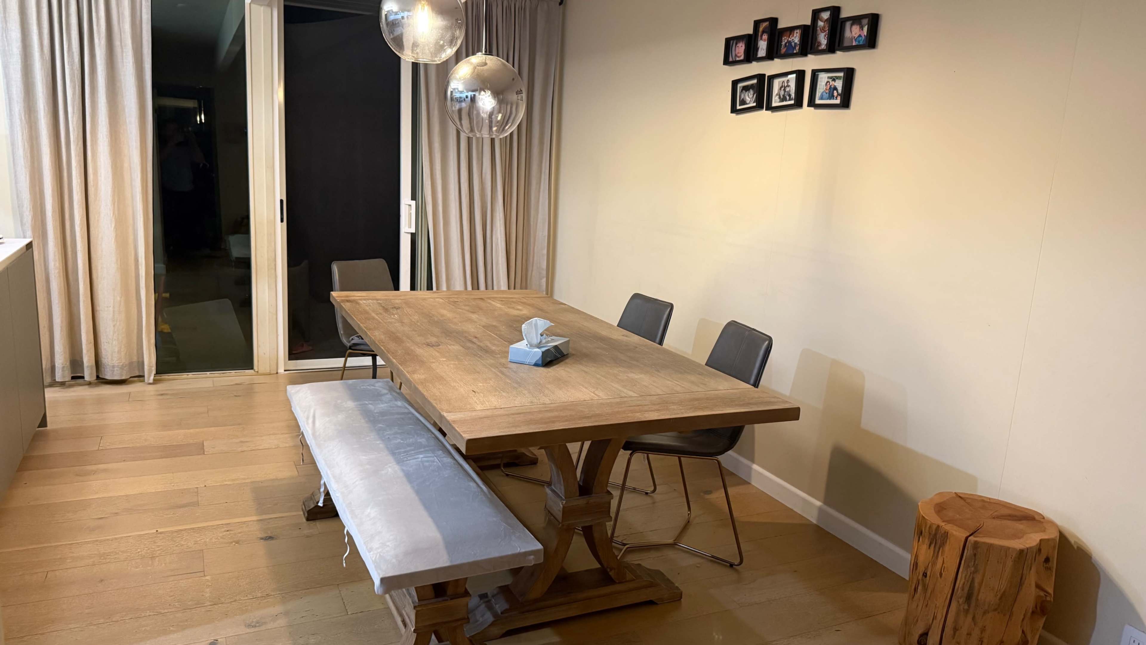A wooden dining table with a bench and chairs is positioned against a wall featuring family photos, with large curtains and a sliding glass door in the background.