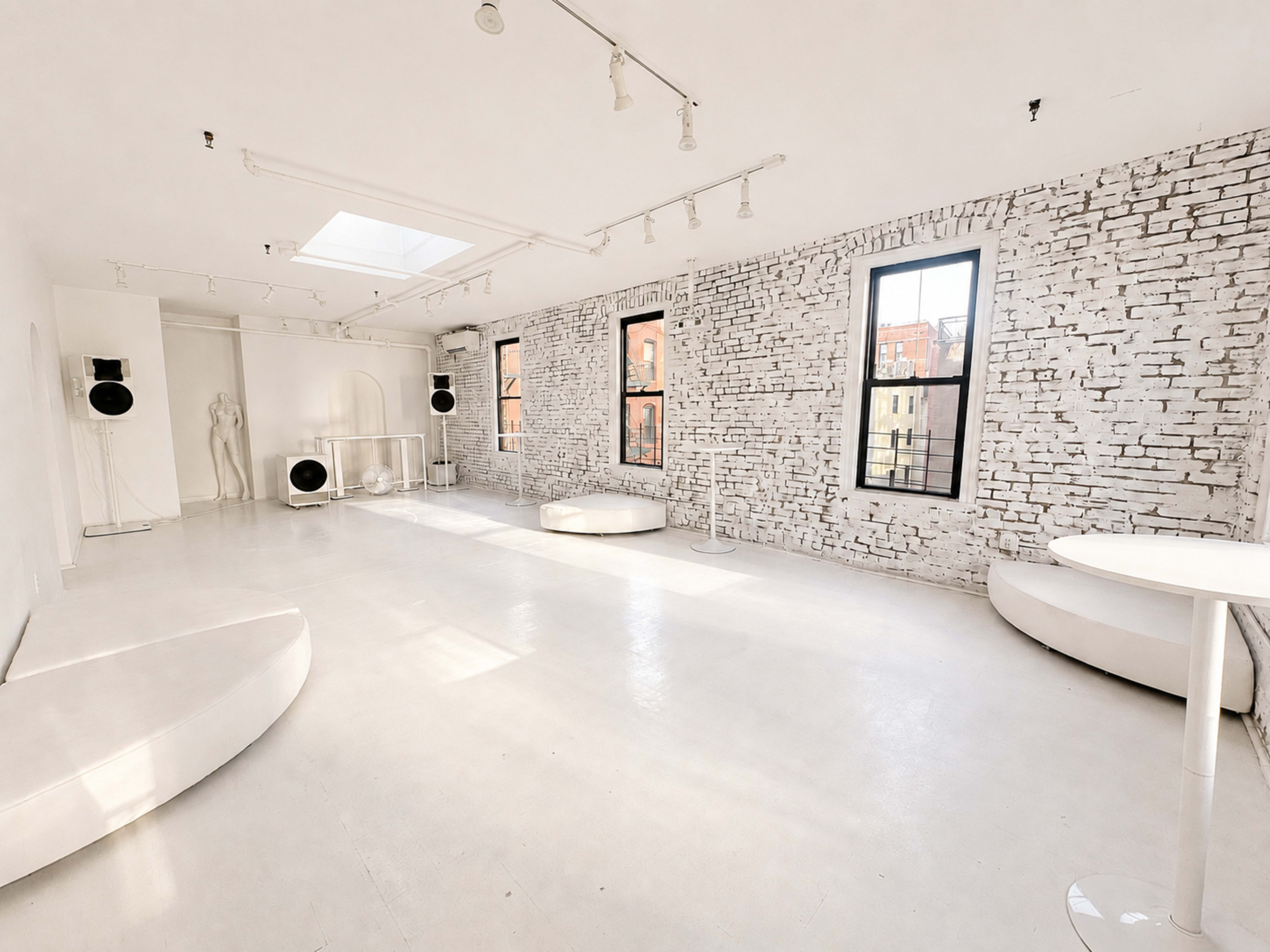 The image shows a spacious, minimalist gallery interior with white walls, a polished floor, and large windows.