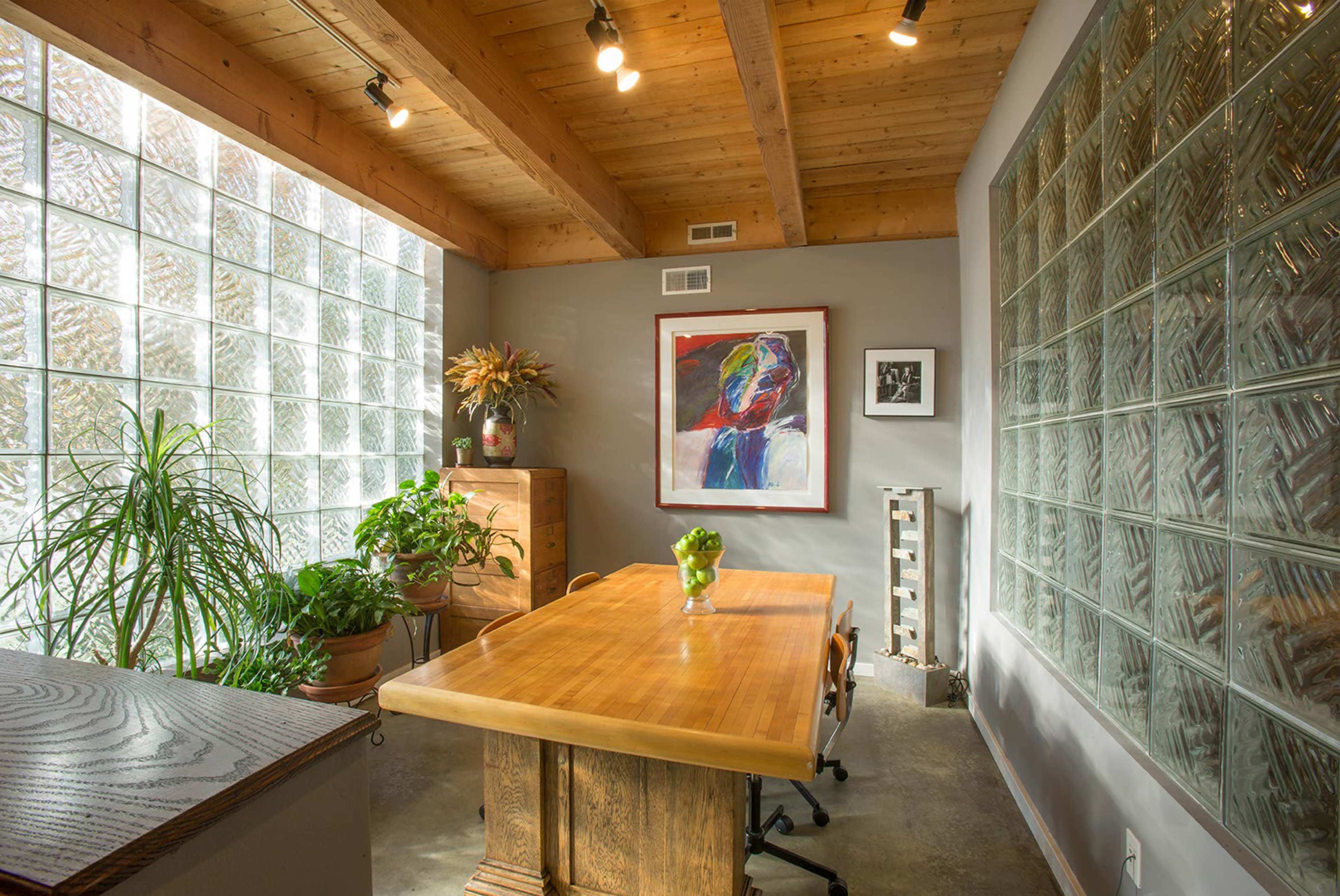 A well-lit office space features a large wooden table surrounded by chairs, with glass block walls and potted plants.