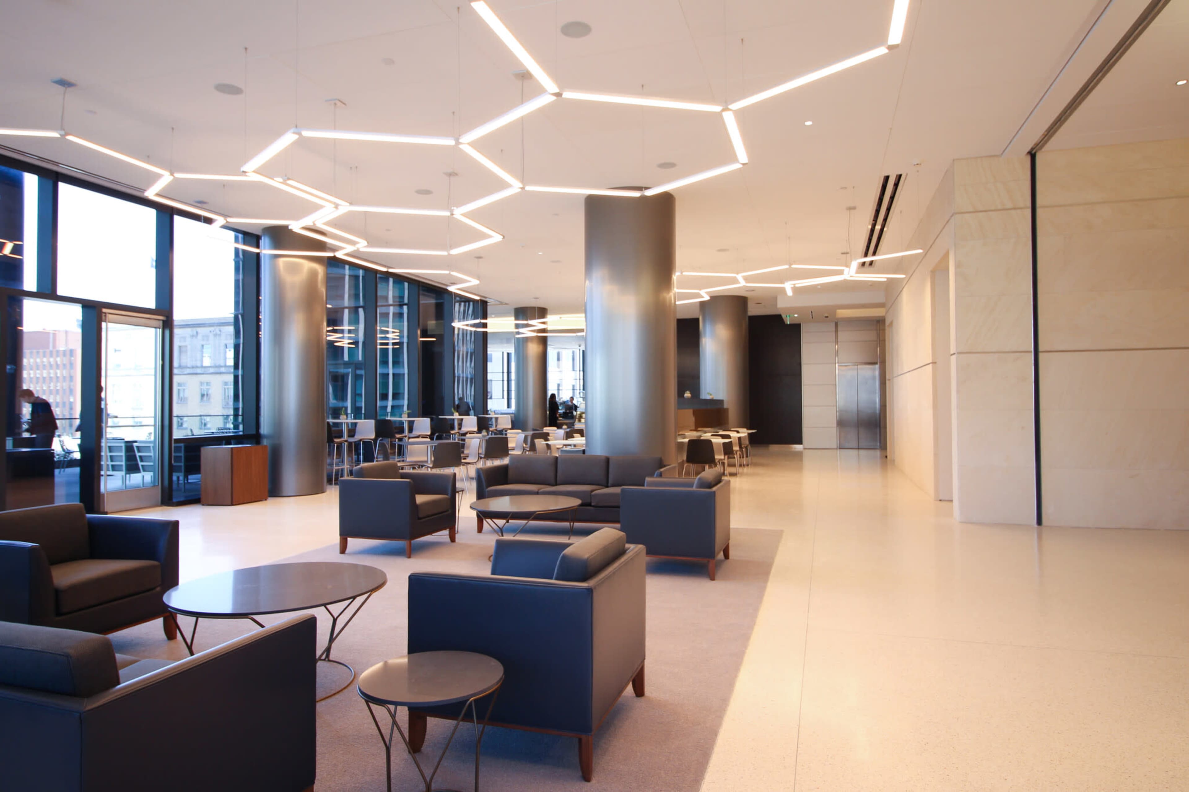 The image shows a modern office lobby with hexagonal light fixtures, seating areas with dark couches, and large windows allowing natural light.