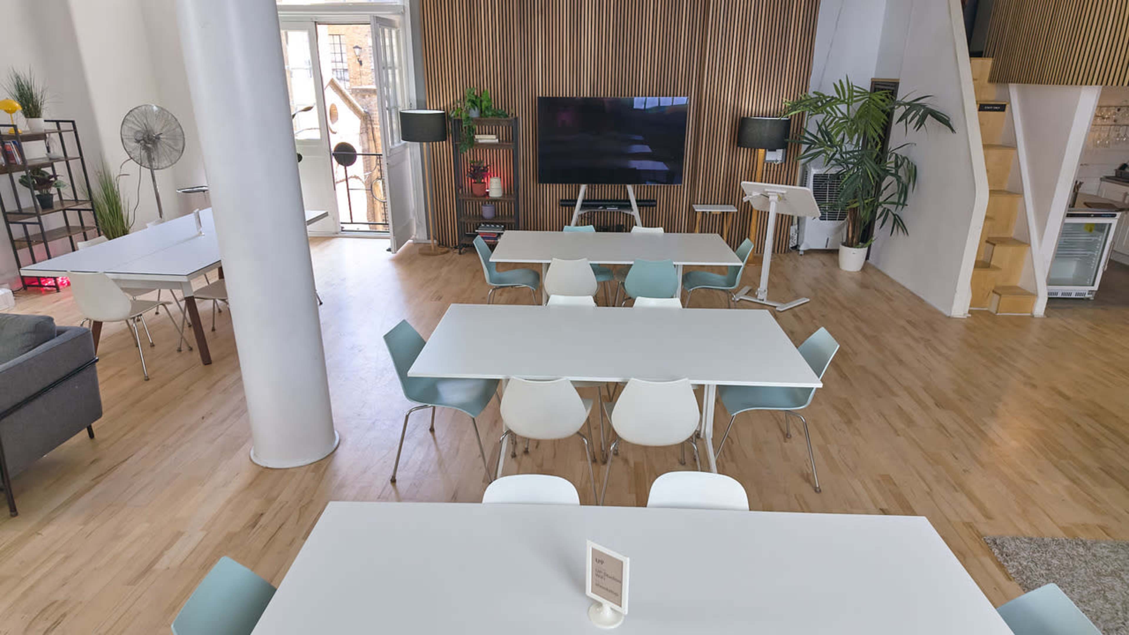 The image shows a spacious, modern interior with multiple white tables, chairs, a large television, and decorative plants.